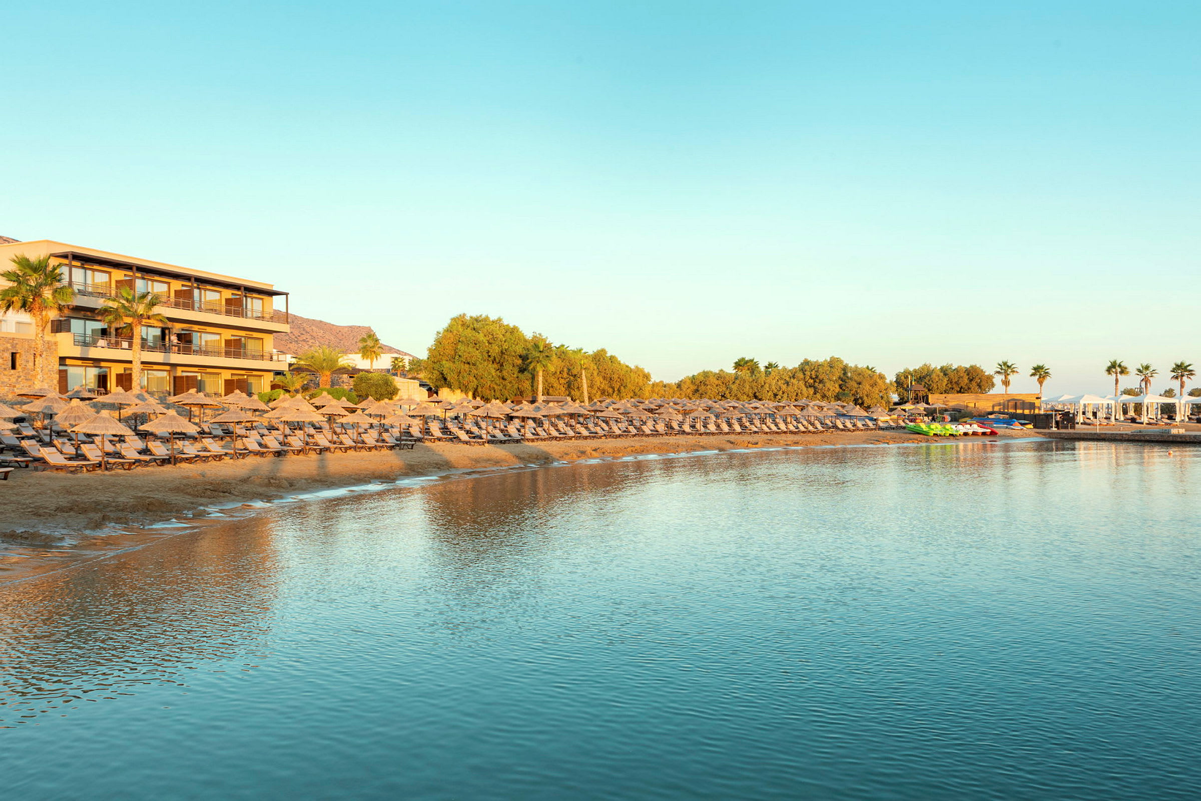 En strand med solsenger og parasoller ved et hotell med utsikt over rolig vann. (AI-generert tekst)