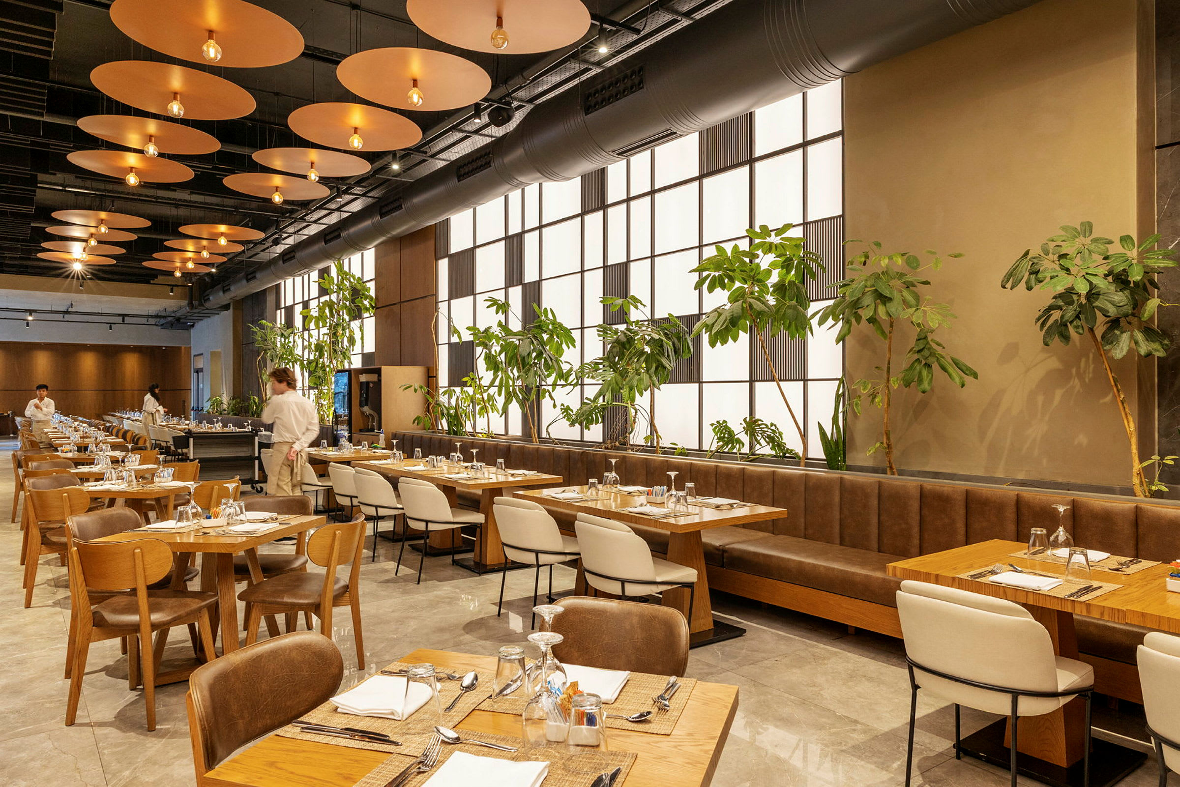 En moderne restaurant med trebord og stoler, grønne planter langs en vegg og en servitør synlig i bakgrunnen. (AI-generert tekst)