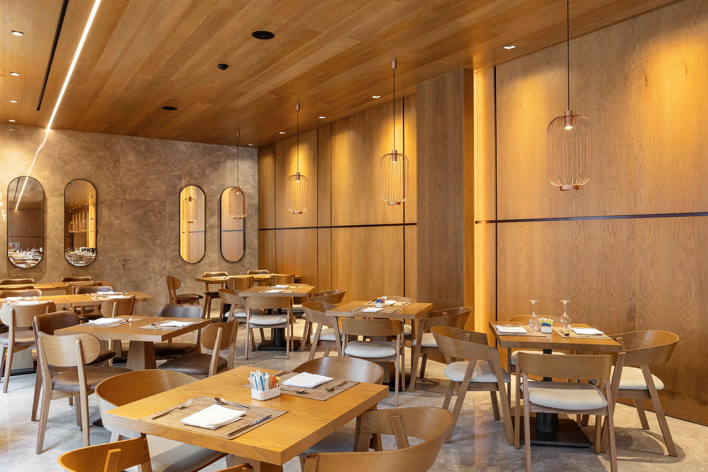 En moderne restaurant med trepaneler på vegger og tak, elegante hengende lamper og dekkede bord med trestoler. (AI-generert tekst)