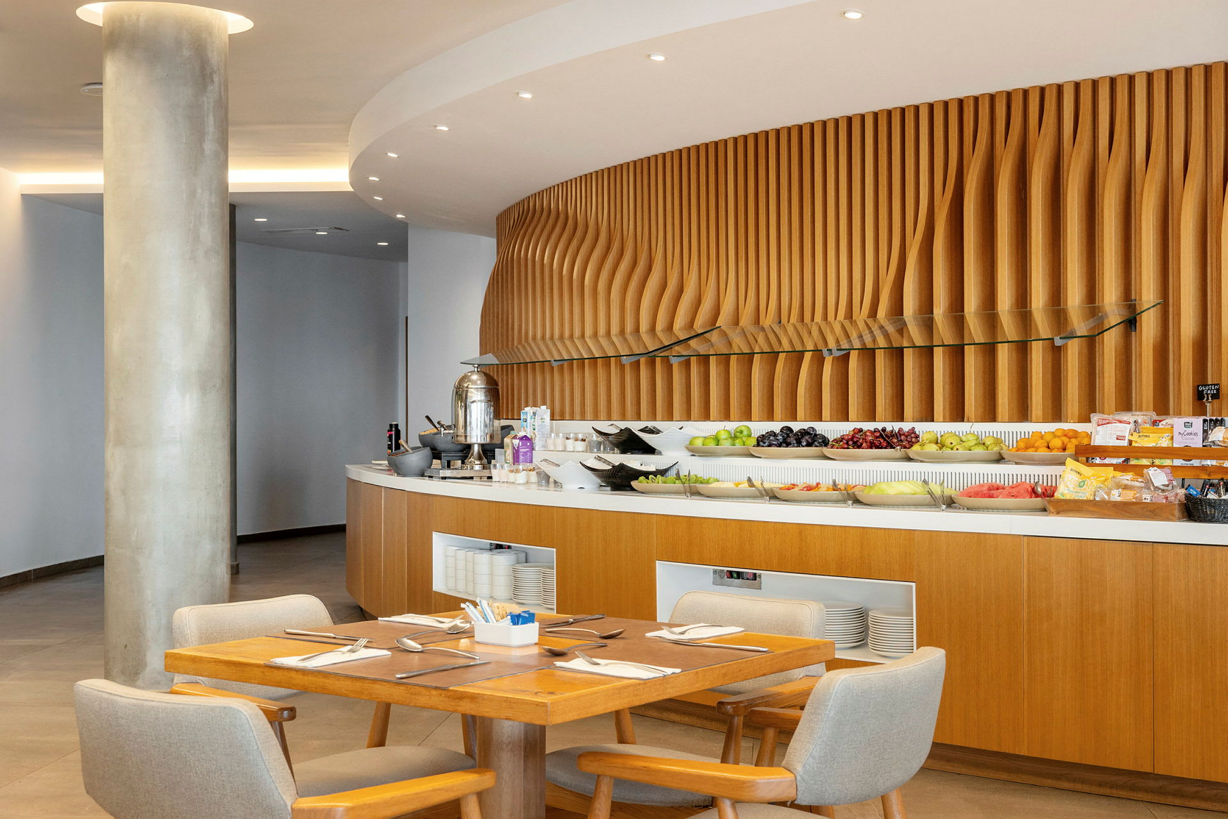 En frokostbuffet med frukt og andre tilbehør plassert på en trebelagt disk med en dekorativ vegg i bakgrunnen. Foran står et bord og stoler. (AI-generert tekst)