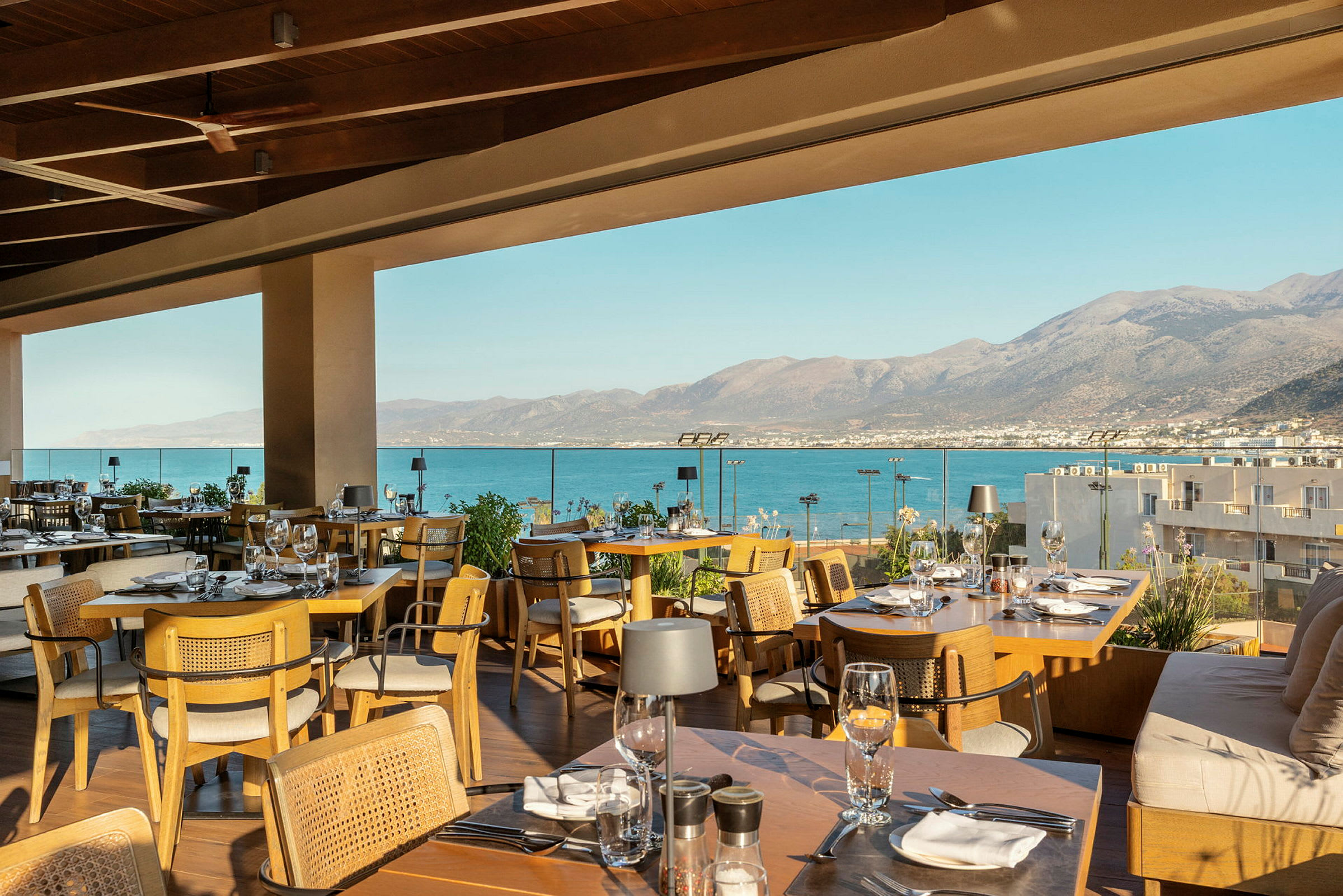 En restaurant med utsikt over havet fra en terrasse. Uteområdet er møblert med trebord og stoler, og fjellene i bakgrunnen strekker seg utover vannet. (AI-generert tekst)