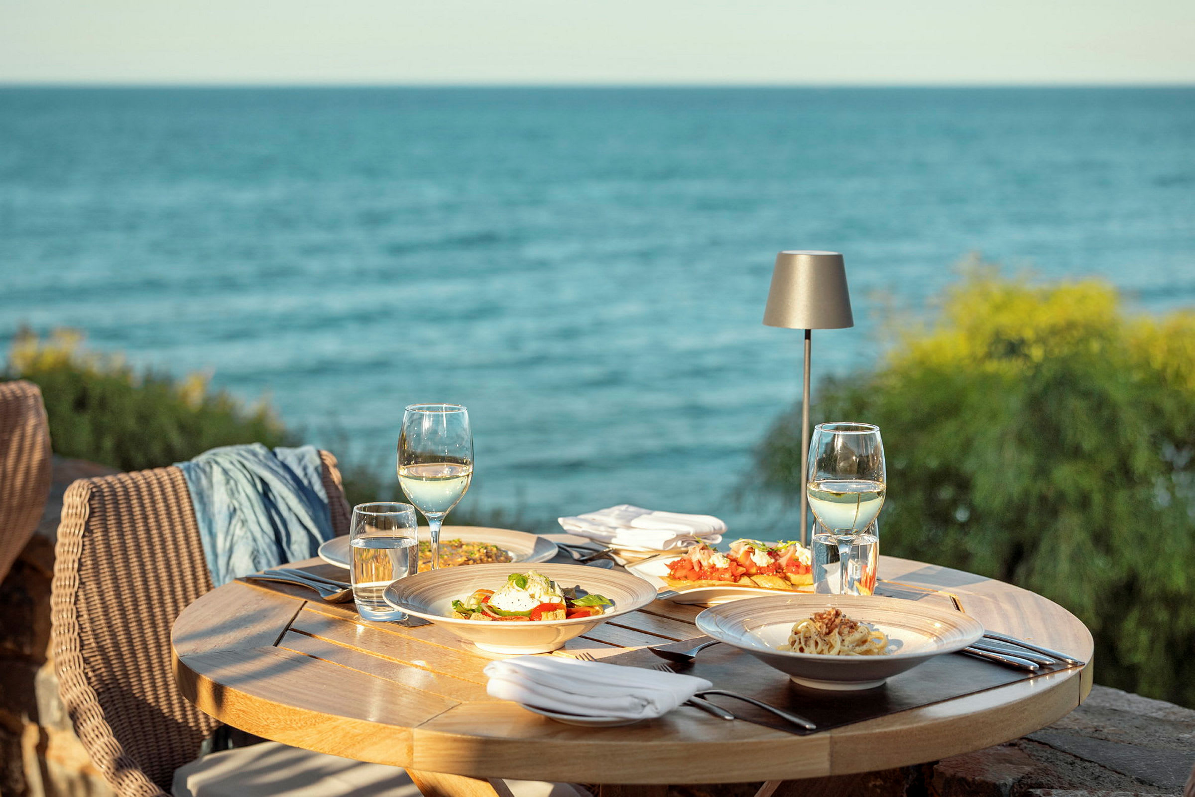 Uteservering ved havet med et rundt bord fullt av tallerkener med mat, vinglass og bestikk. (AI-generert tekst)