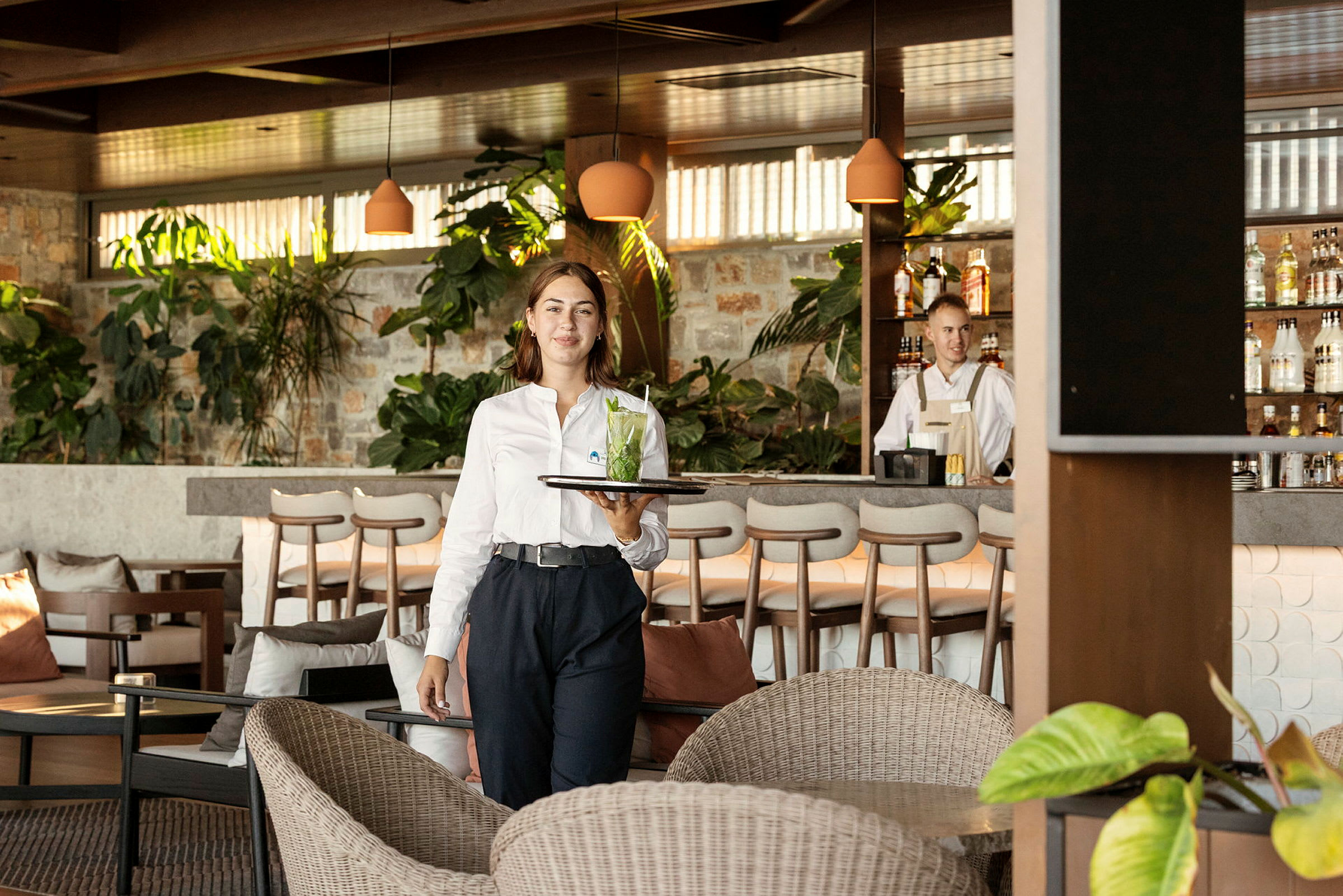 En servitør bærer et brett med en drink i en hyggelig lounge med planter og en bardisk i bakgrunnen. (AI-generert tekst)