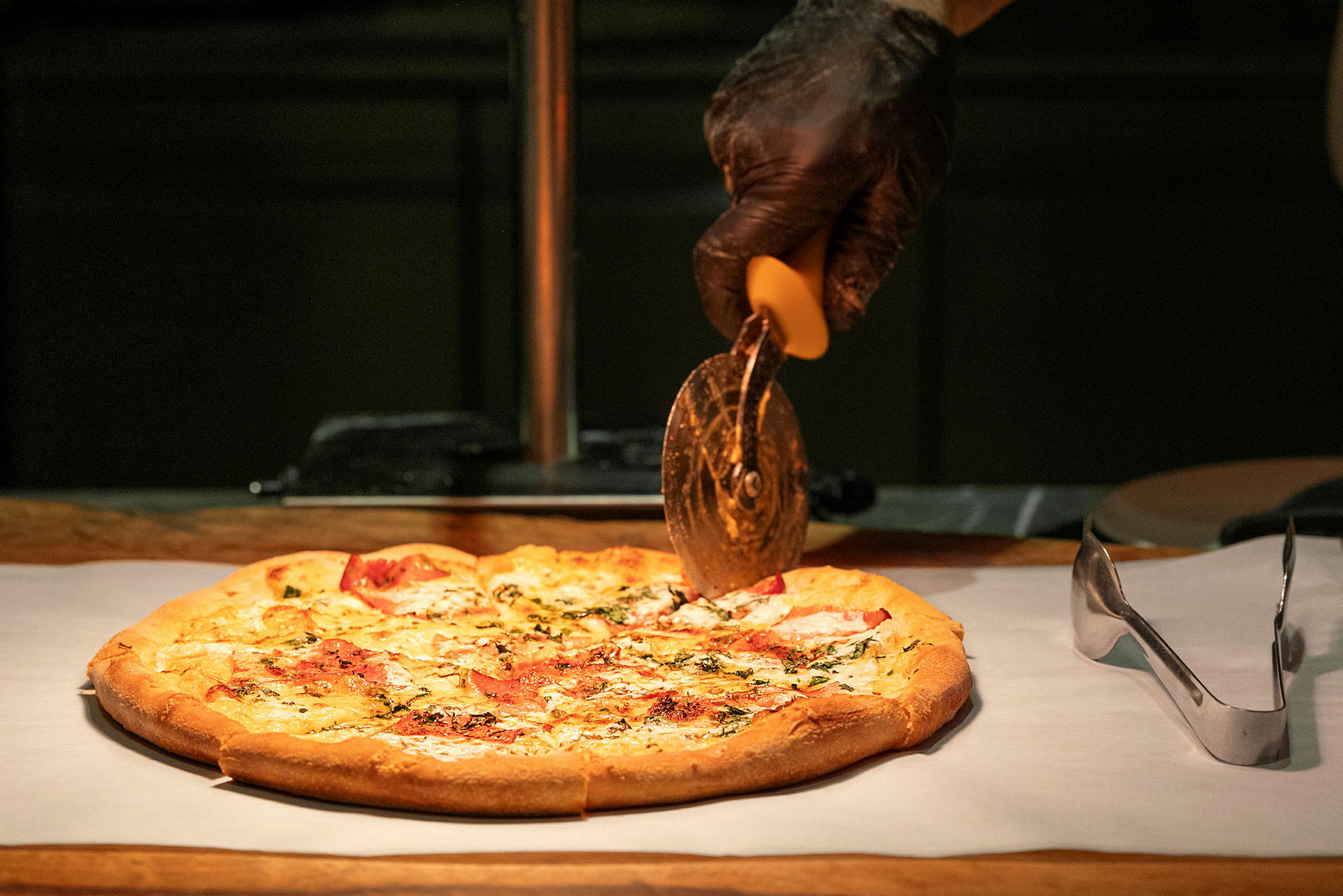 En pizza på et bakepapir skjæres med en pizzaskjærer som holdes av en hånd i svart hanske. (AI-generert tekst)