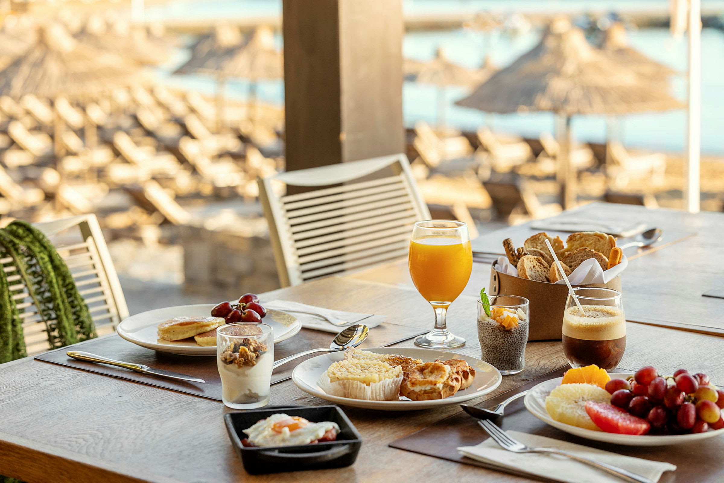 Et bord med en frokostoppsetning, inkludert glass med juice og kaffe, bakverk, frukt og yoghurt. I bakgrunnen ses parasoller på stranden. (AI-generert tekst)