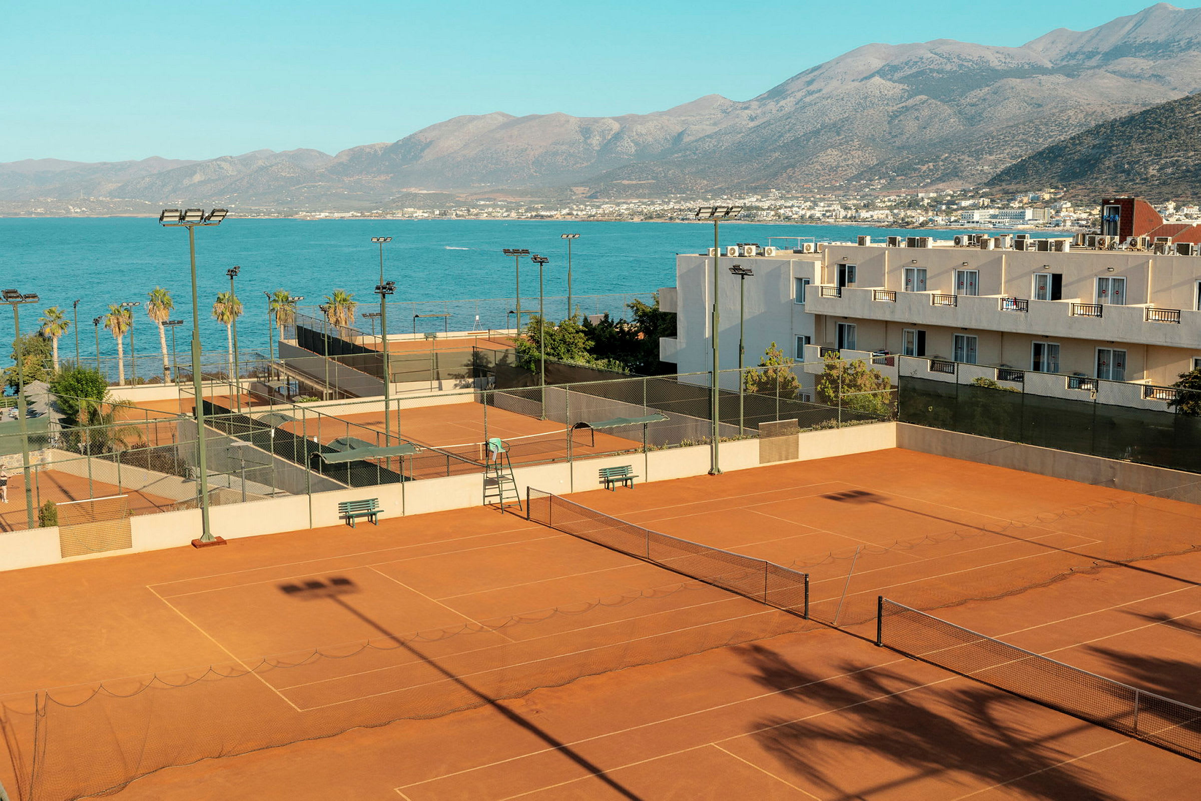 Et bilde av flere tennisbaner laget av grus, plassert ved kysten nær havet med fjell i bakgrunnen. (AI-generert tekst)