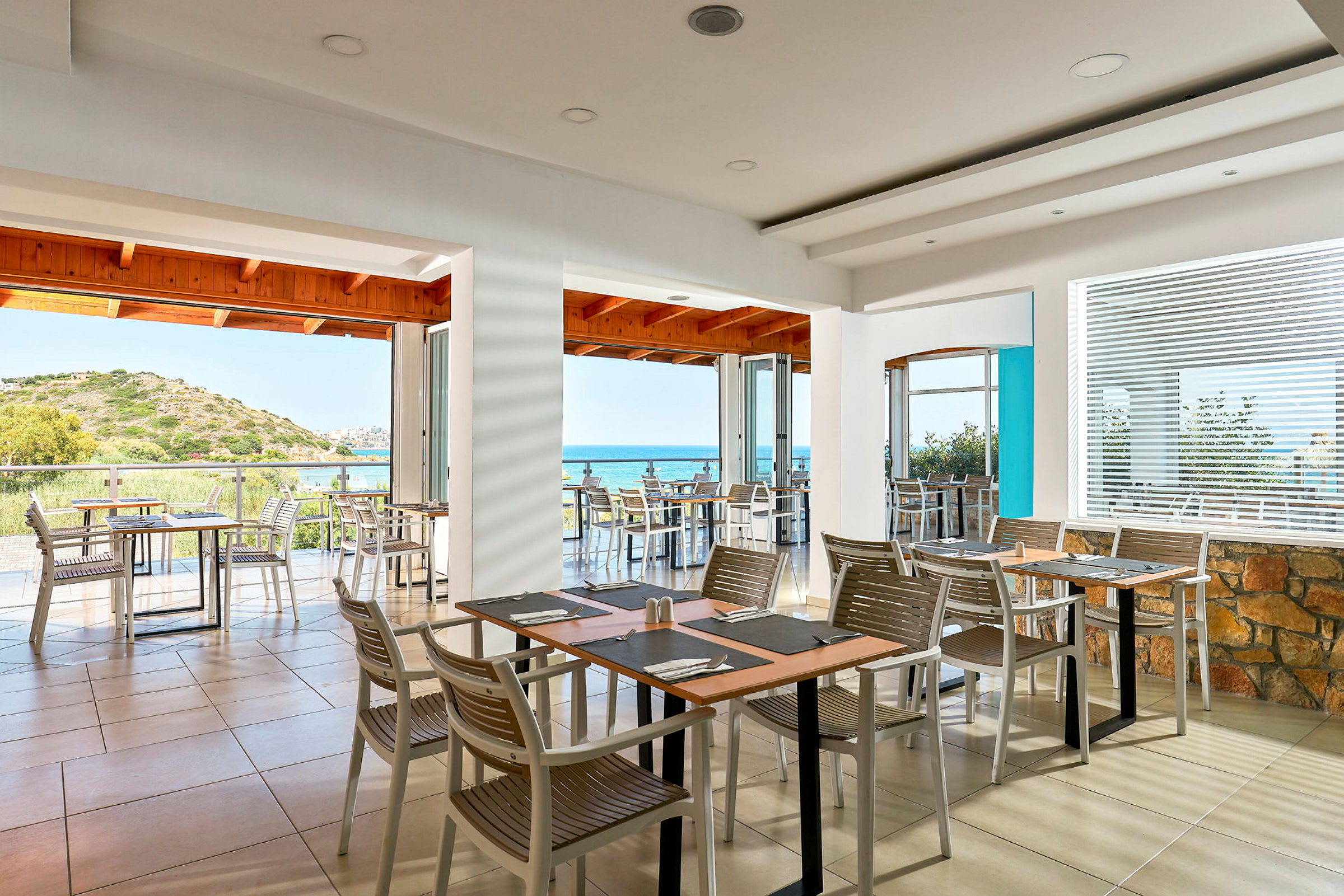 En restaurant med utsikt over havet, med bord og stoler innendørs og på en terrasse. (AI-generert tekst)