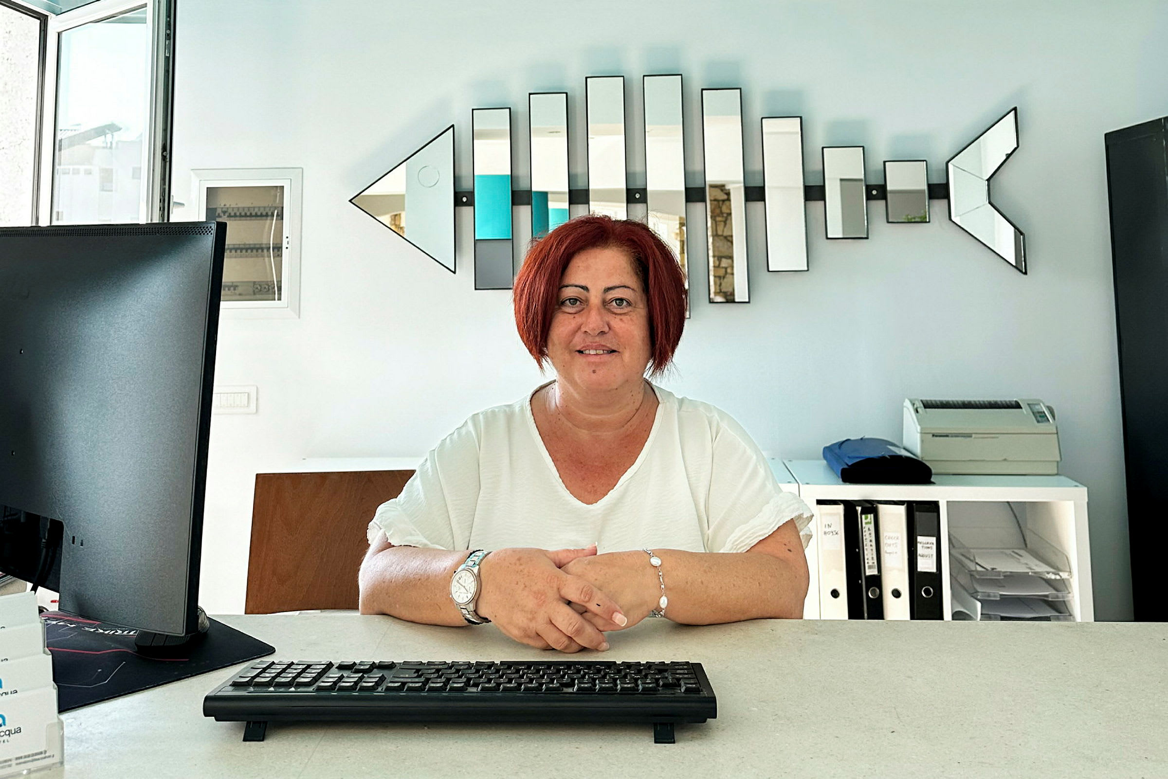 Person sitter ved et skrivebord med tastatur, dataskjerm og hyller i bakgrunnen. (AI-generert tekst)