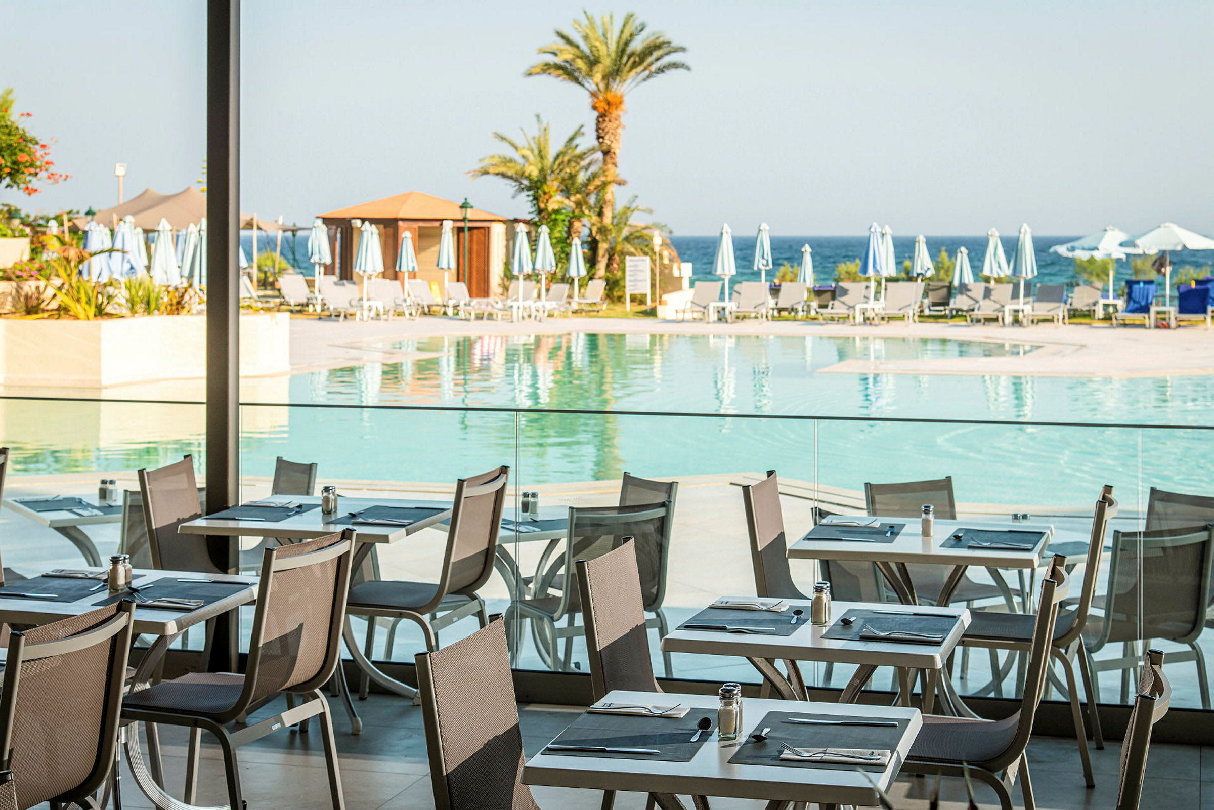Motiv av et utendørsområde med basseng ved en restaurant med bord og stoler, omgitt av solsenger og parasoller. Palmer ses, og havet er i bakgrunnen. (AI-generert tekst)