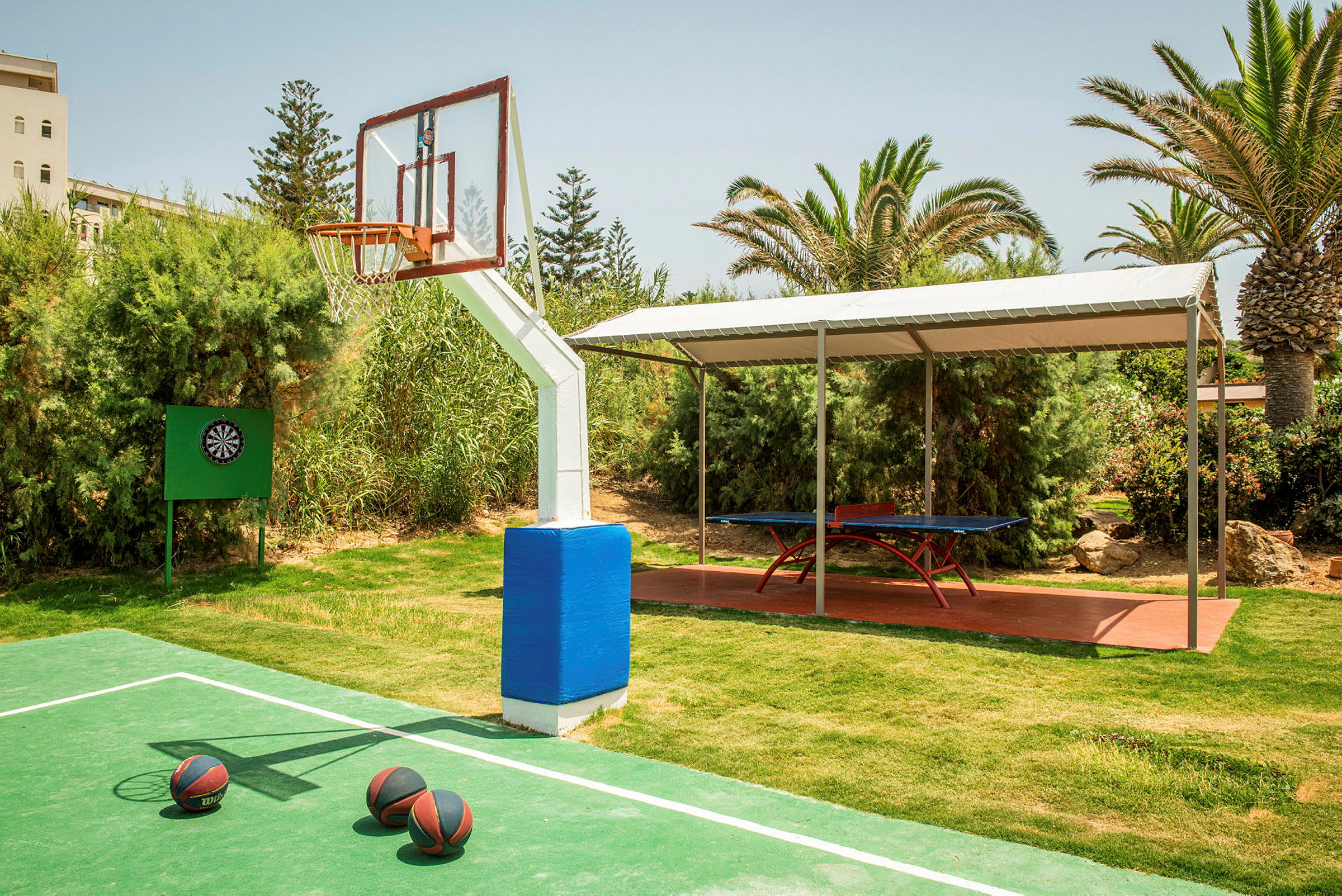 Utendørsområde med basketballbane, darttavle og et bordtennisbord under et tak. (AI-generert tekst)