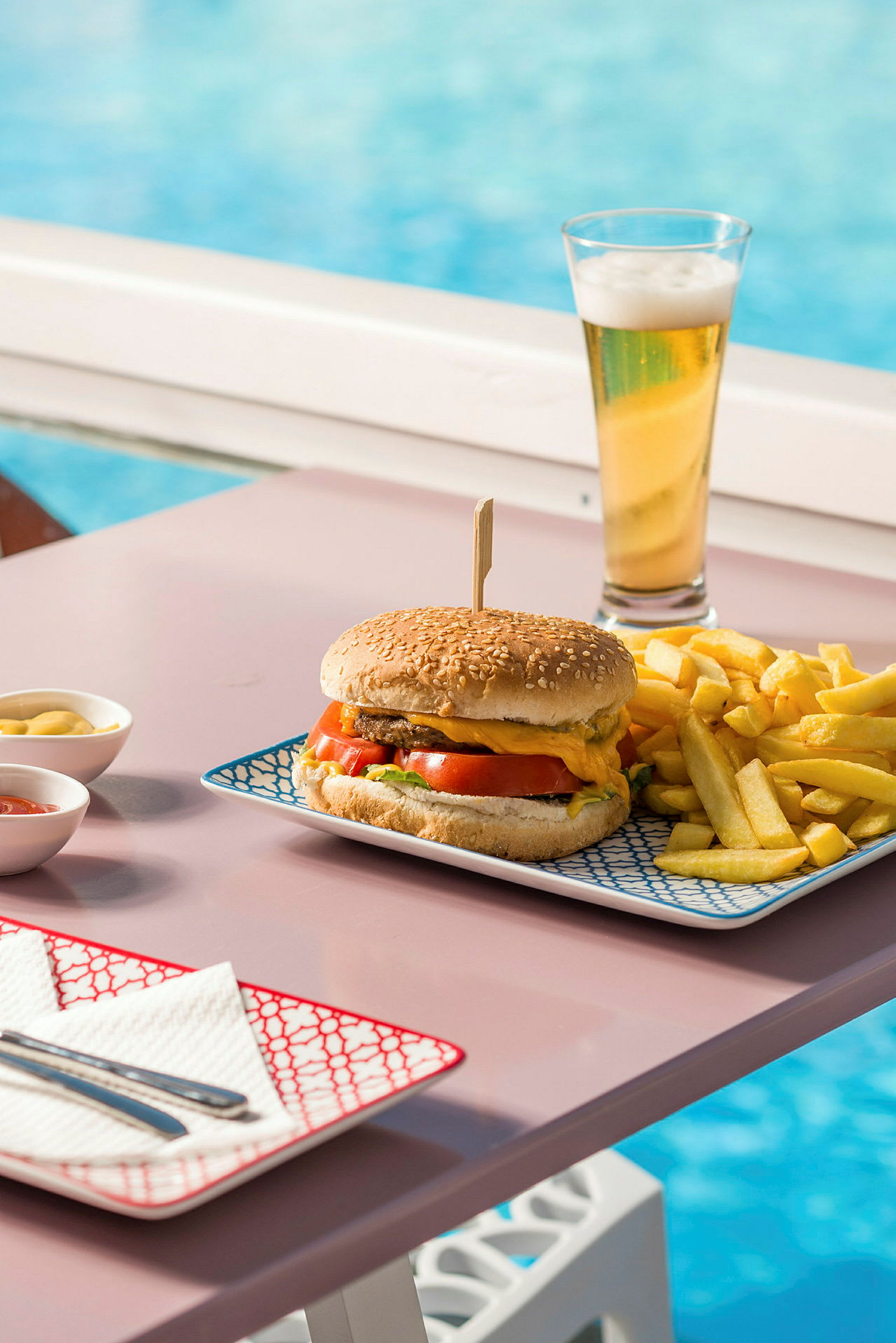 En hamburgerrett med pommes frites og en øl ved siden av et basseng. (AI-generert tekst)