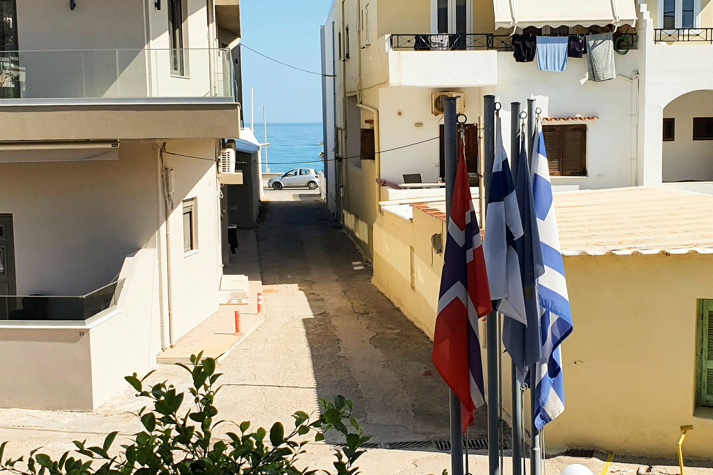 Utsikt over en liten gate med bygninger på begge sider og havet synlig i bakgrunnen. Flere flagg er plassert i forgrunnen. (AI-generert tekst)