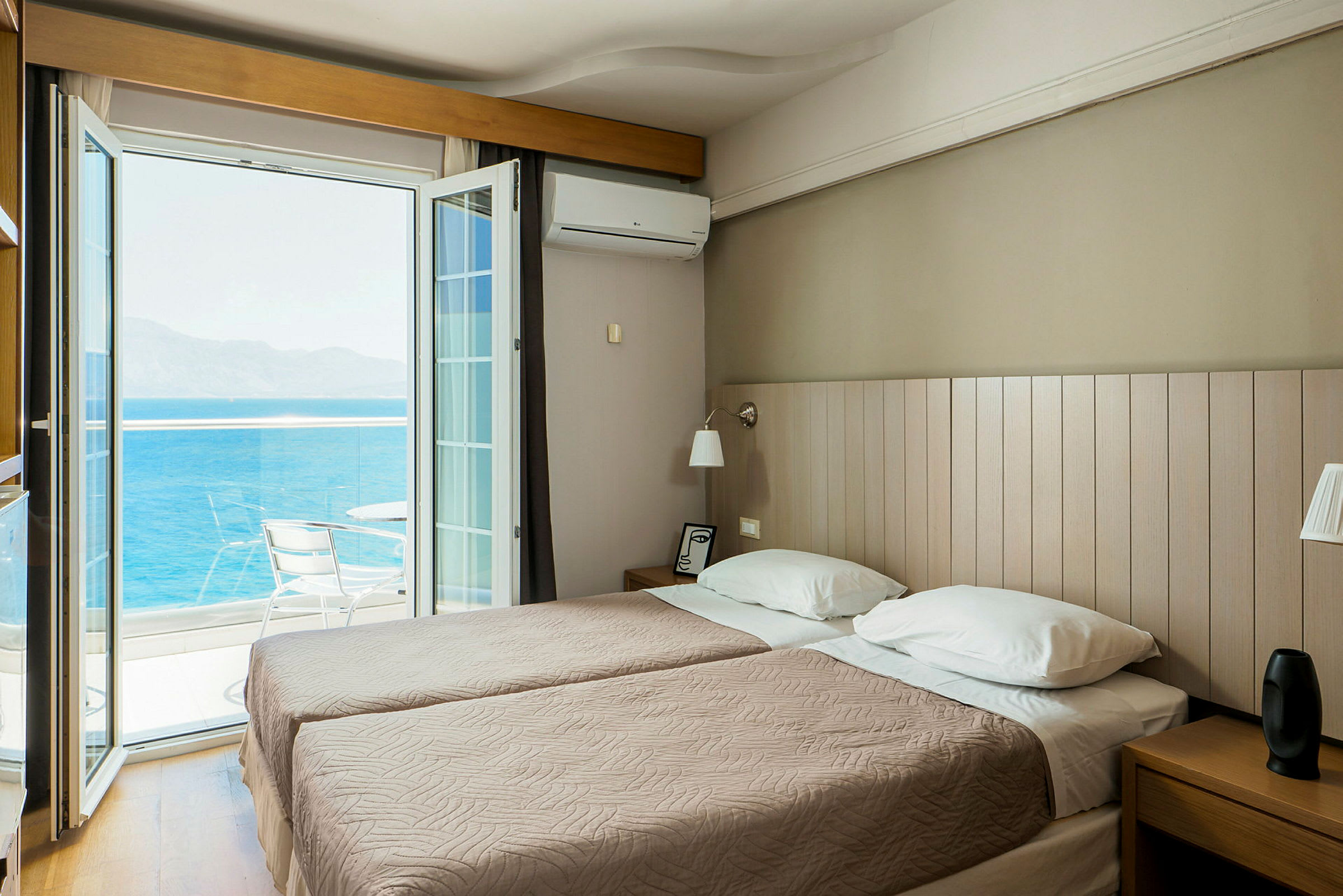 Et hotellrom med to enkeltsenger og en åpen balkongdør som vender mot havet. (AI-generert tekst)