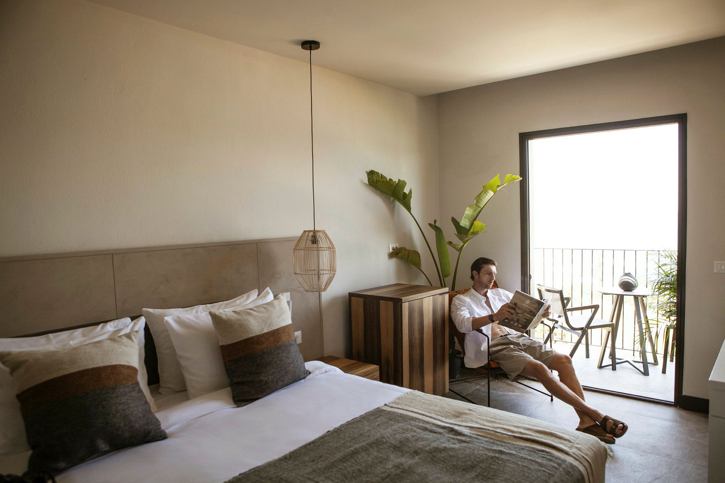 Et hotellrom med en oppredd dobbeltseng, moderne interiør, og en balkong med utemøbler samt grønt. (AI-generert tekst)