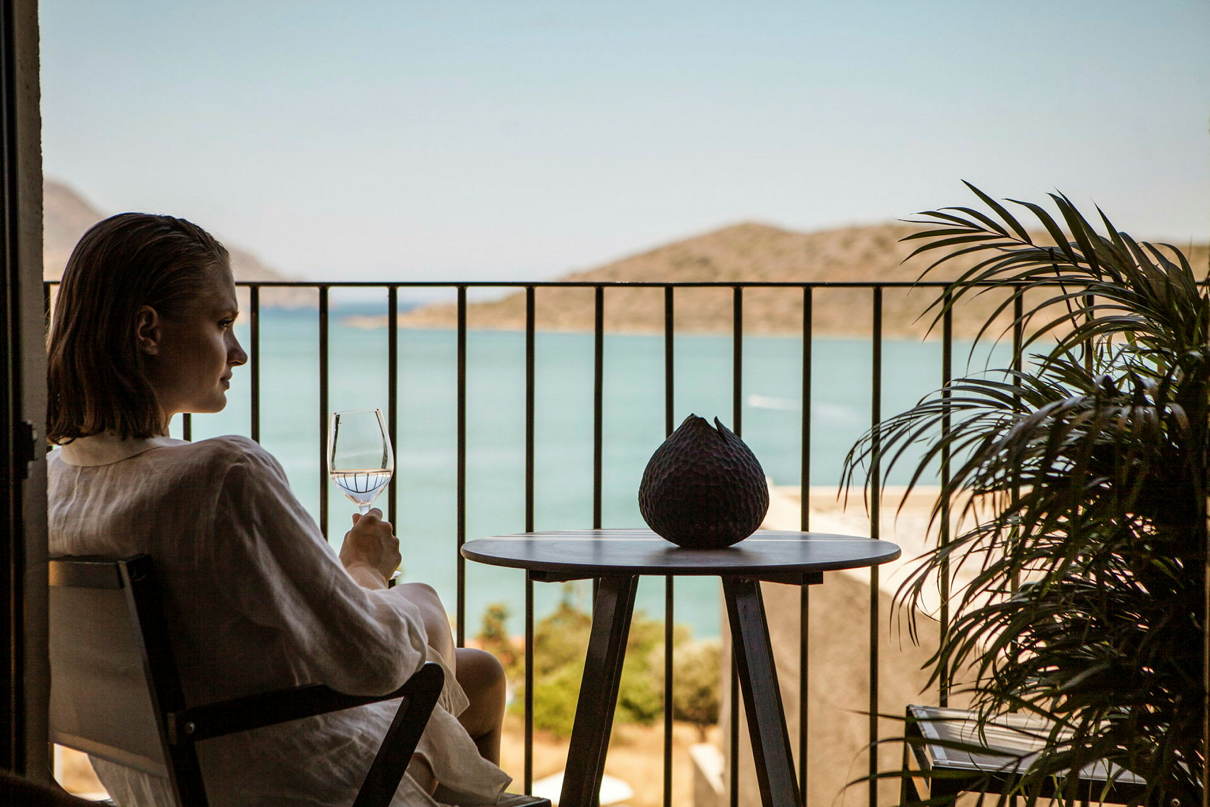 En person sitter på en balkong og holder et glass med utsikt over havet. (AI-generert tekst)