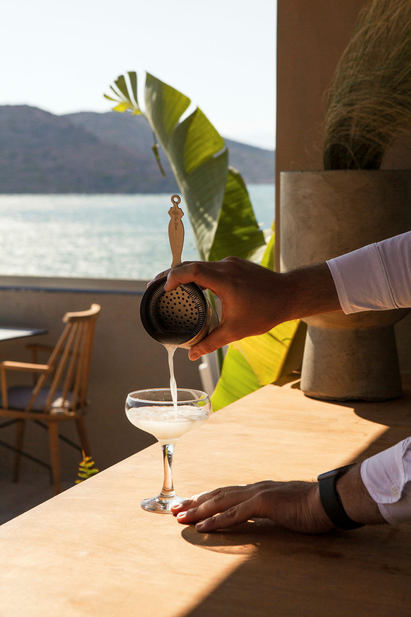 En hånd som heller en drikke gjennom en sil i et cocktailglass, med havutsikt og planter i bakgrunnen. (AI-generert tekst)