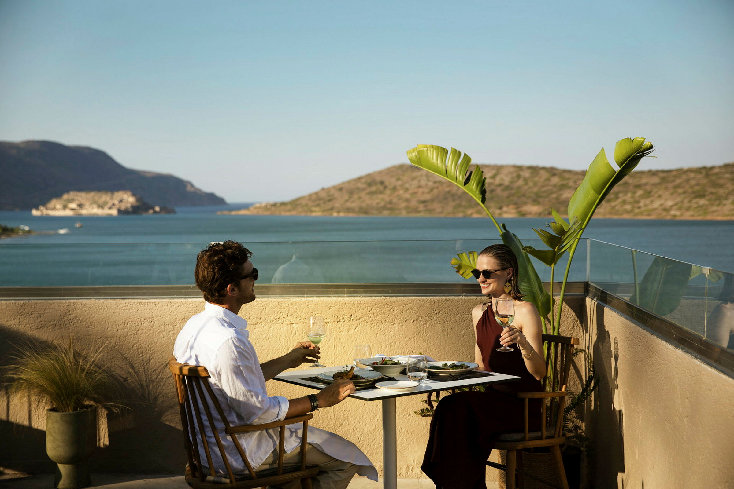 To personer sitter ved et bord med mat og drikke på en terrasse med utsikt over havet og fjell. (AI-generert tekst)