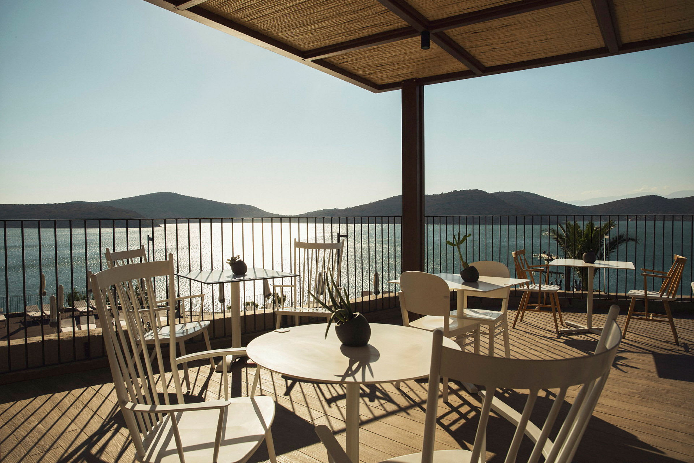 Terrasse med bord og stoler, utsikt over hav og fjell i bakgrunnen. (AI-generert tekst)