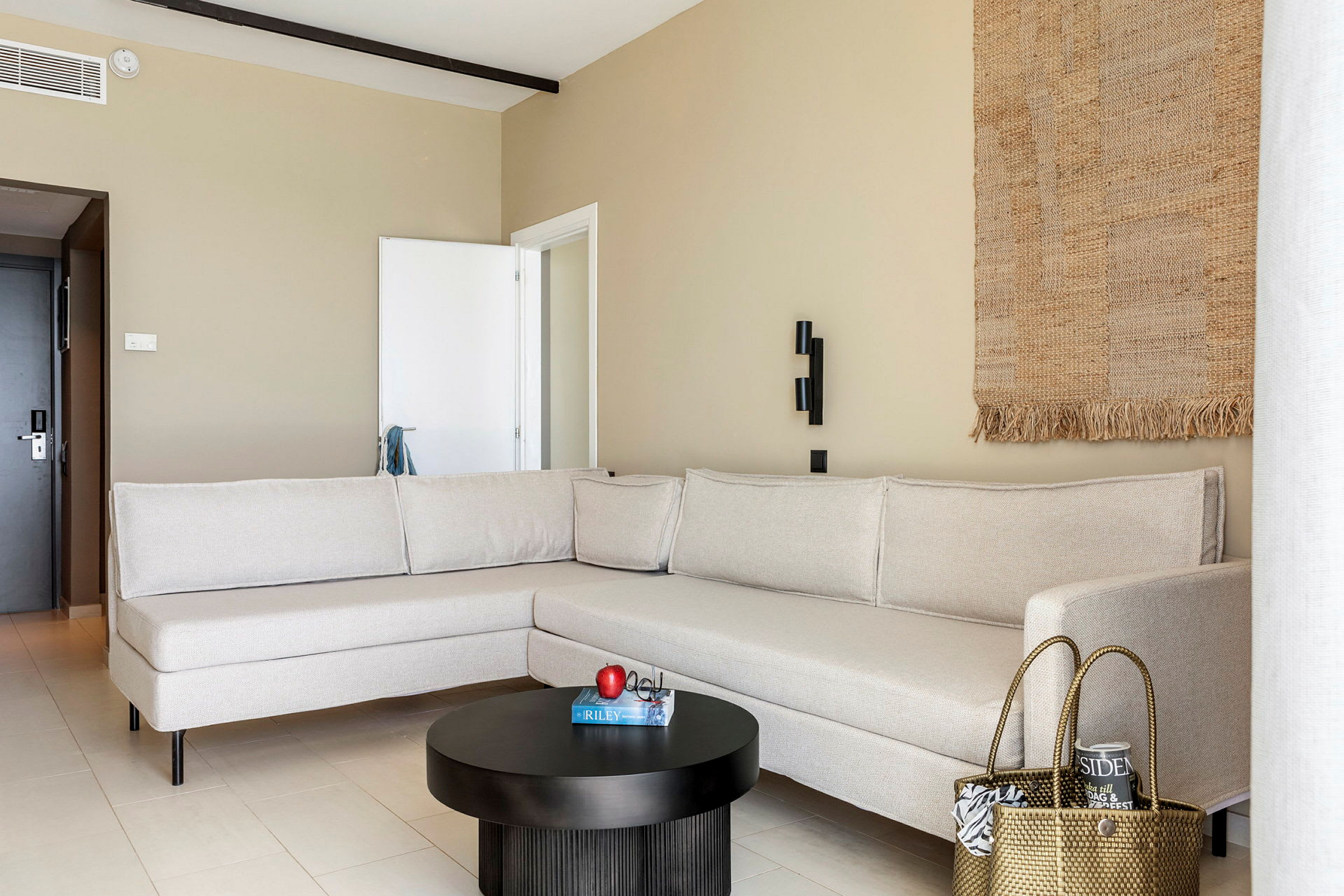 En moderne stue med en beige hjørnesofa, et svart rundt salongbord med et eple og en bok på toppen, og et vevd dekorativt veggteppe. (AI-generert tekst)