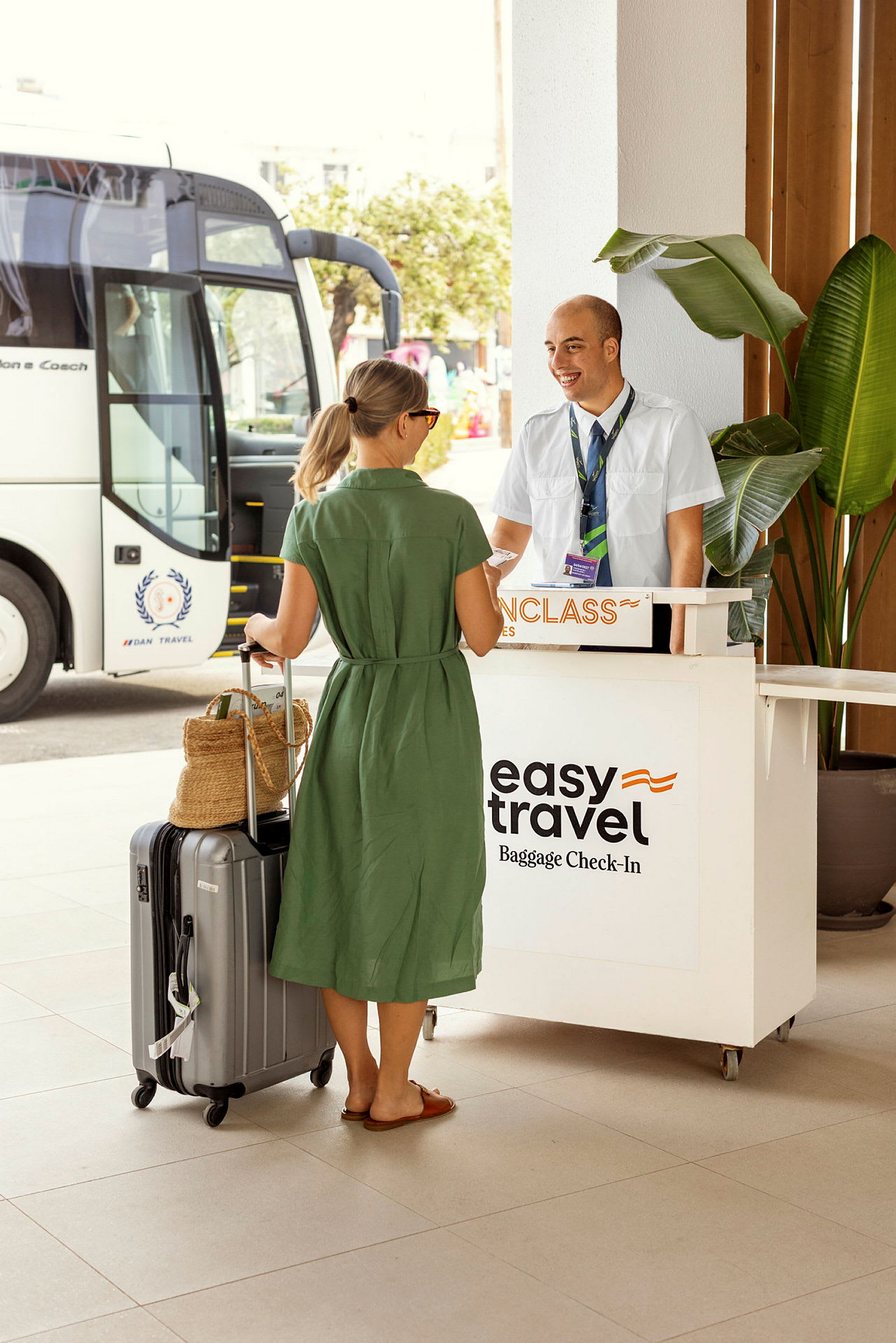 En kvinne står ved en resepsjonsdisk med en koffert og en bag, nær en turbuss. Skiltet på disken indikerer bagasjeinnsjekking. (AI-generert tekst)