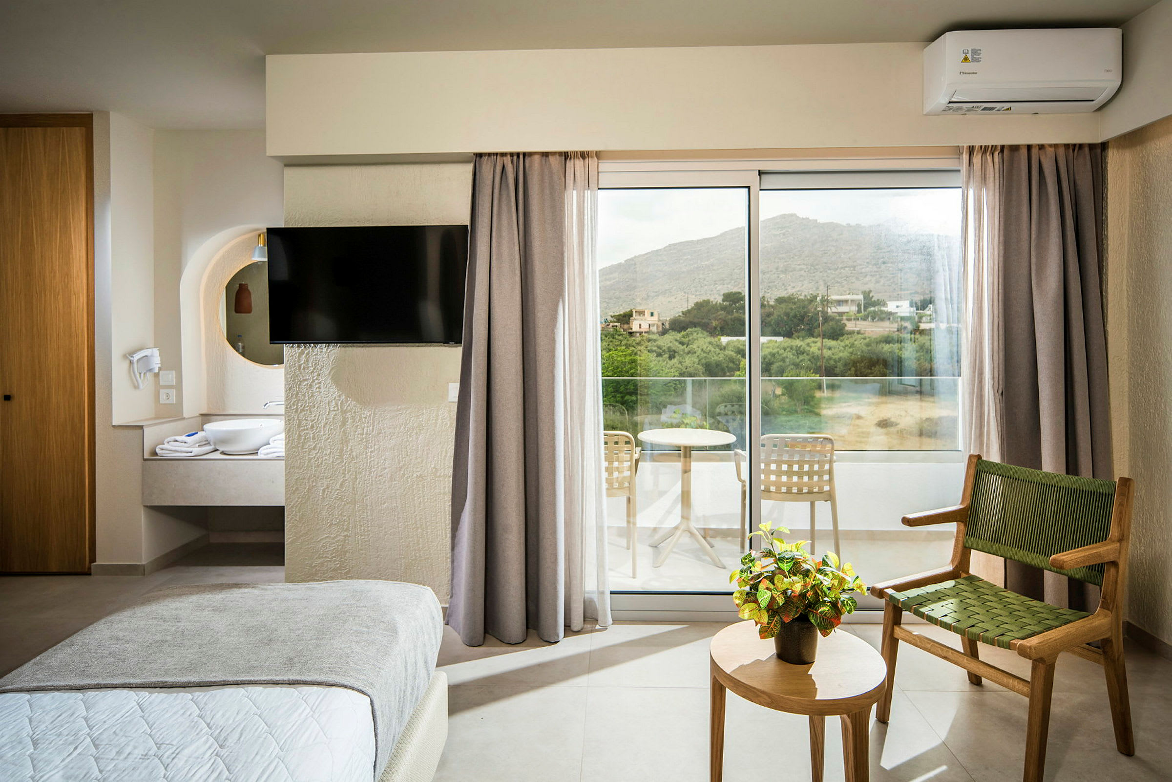 Hotellrom med seng, TV, stol, bord med en plante og utgang til balkong. (AI-generert tekst)