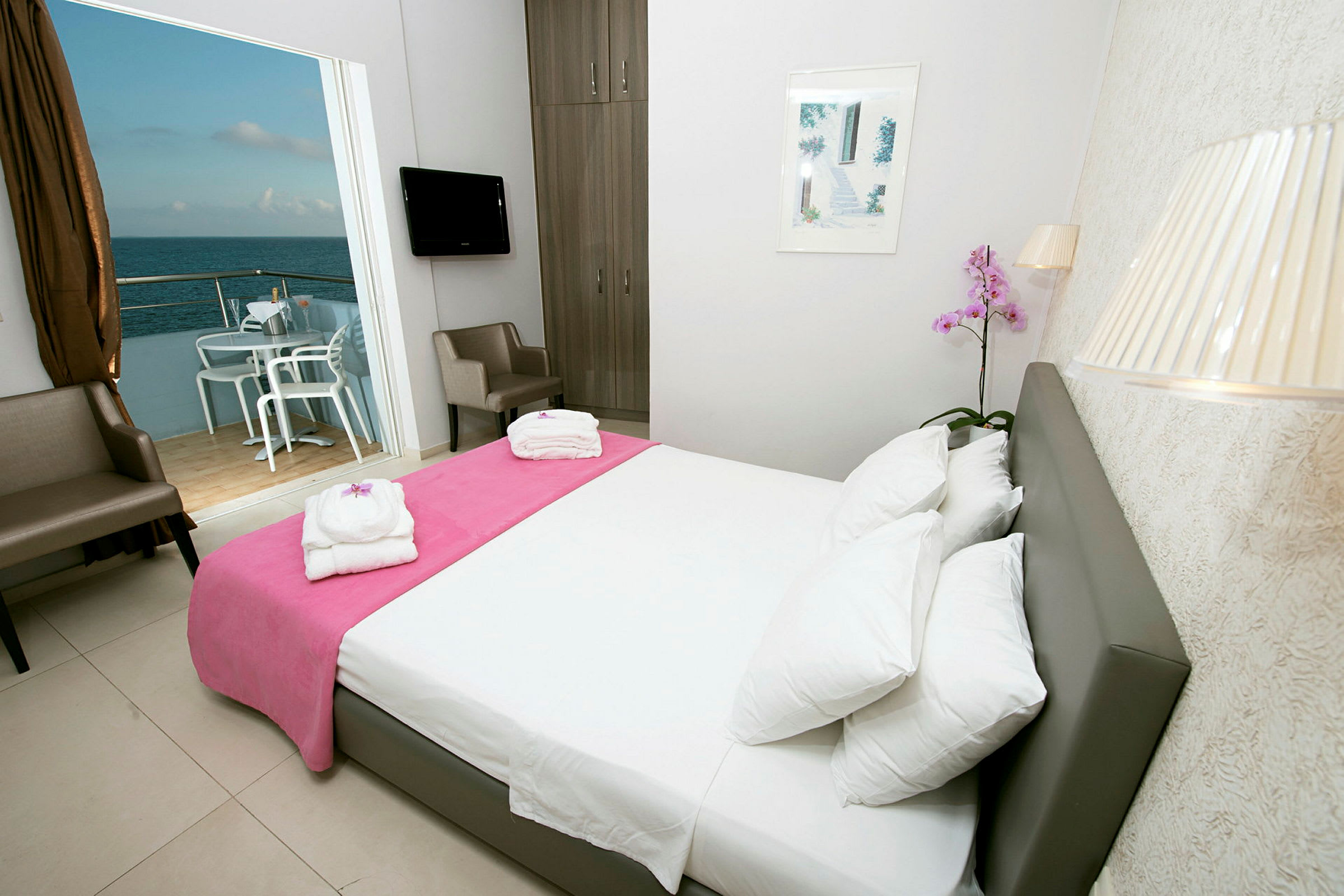 Et hotellrom med hvite vegger, en dobbeltseng med en rosa pute, to stoler, en TV og en balkong med utsikt over havet. (AI-generert tekst)