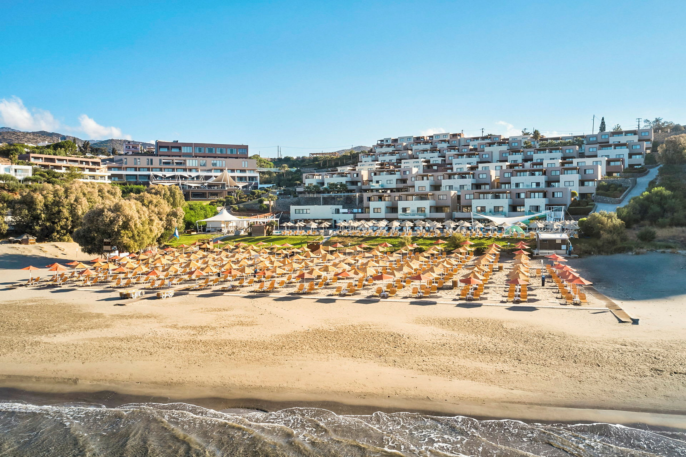En sandstrand med solsenger og parasoller foran en resort med flere bygninger. (AI-generert tekst)