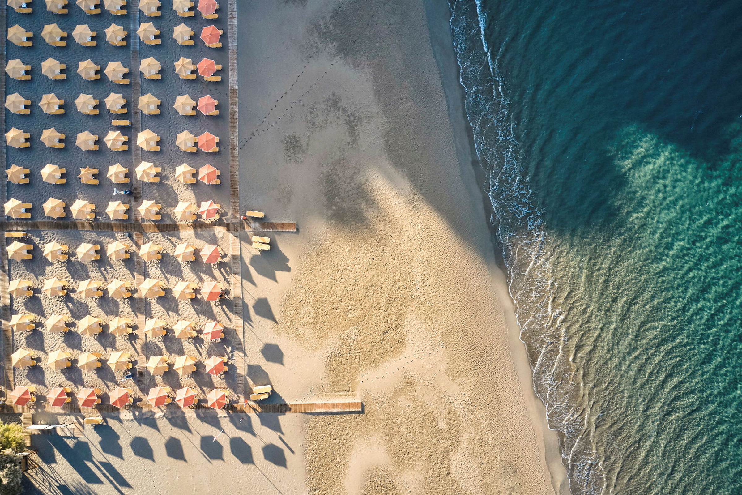Flyfoto av en strand med rader av parasoller og solsenger samt bølger som ruller inn mot sanden. (AI-generert tekst)