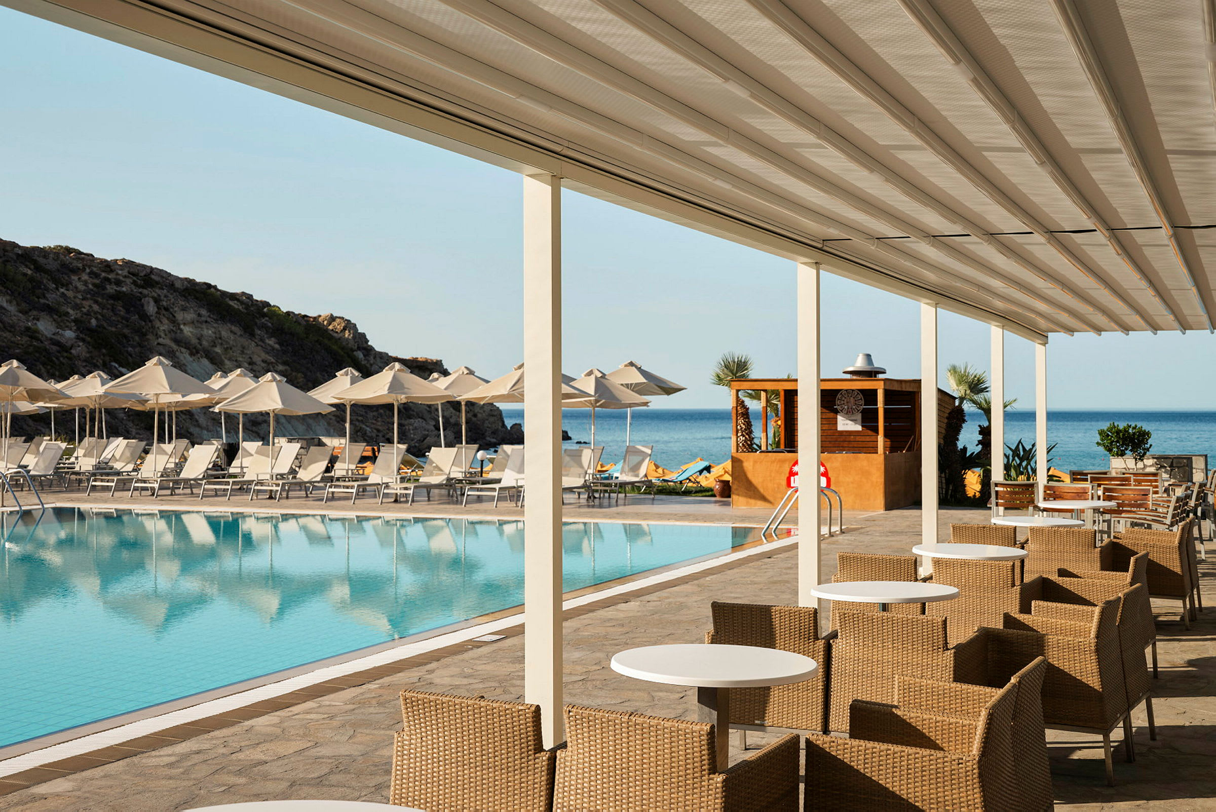 Bilde av et bassengområde ved en resort, med solsenger og parasoller foran havet. Foran bassenget er det sitteplasser med bord og stoler, under en overbygning. (AI-generert tekst)