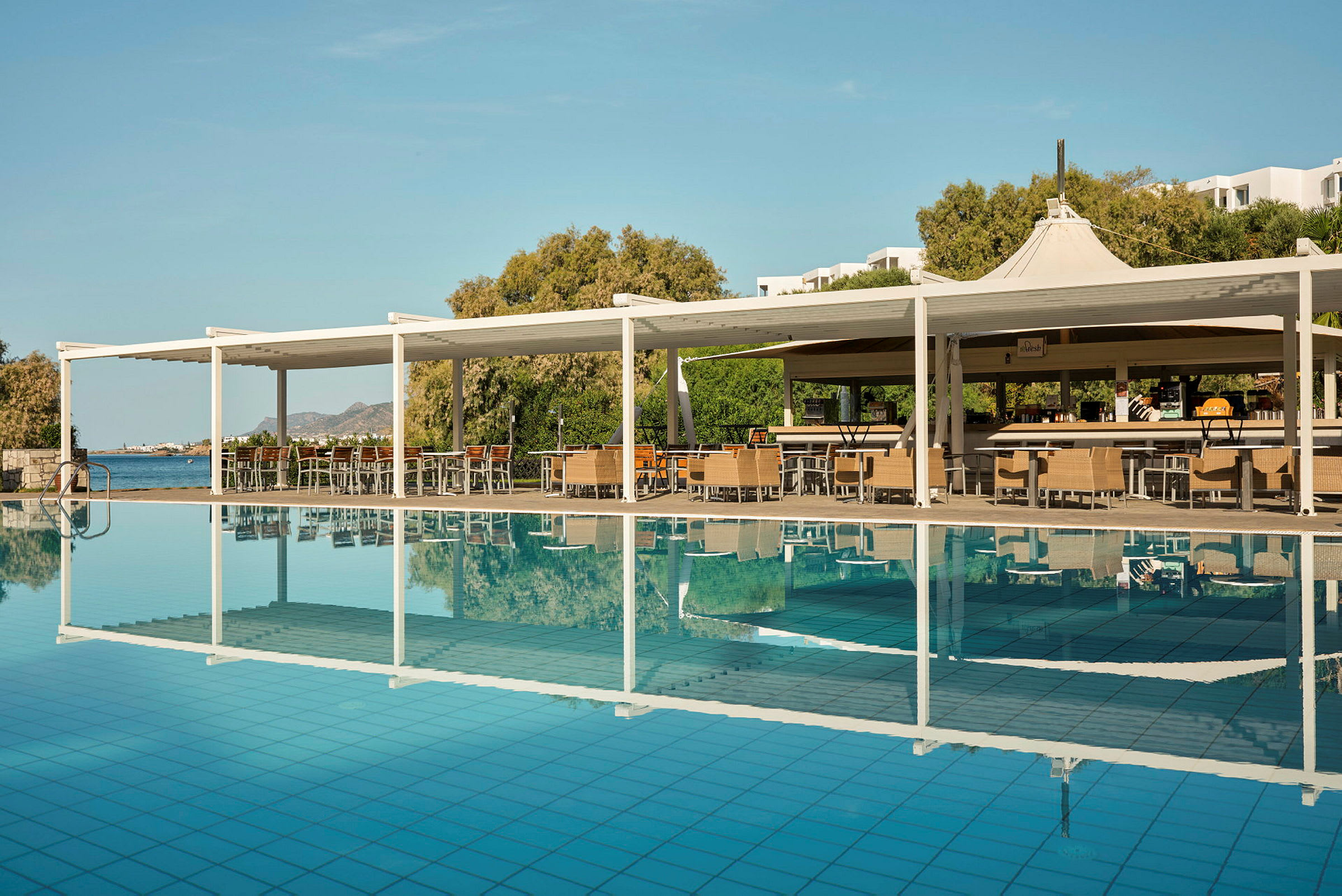 Bilde av et basseng med en tilhørende sitteplass og bar under en hvit pergola. I bakgrunnen sees frodige trær og havet. (AI-generert tekst)