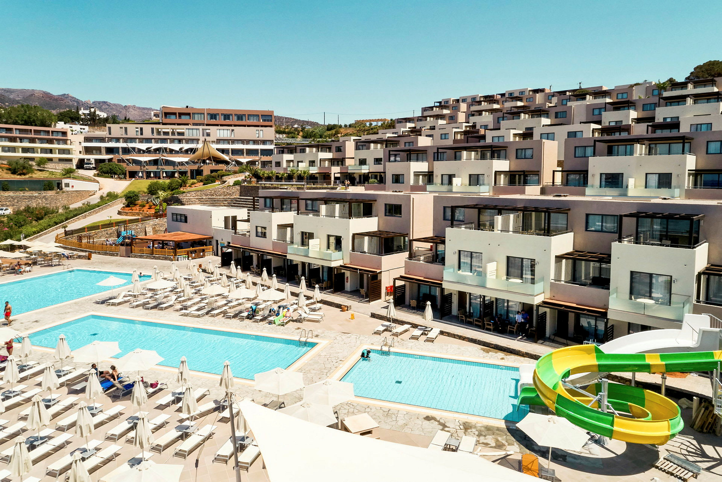 Hotellområde med bassenger, solsenger og vannsklie foran bygninger. (AI-generert tekst)