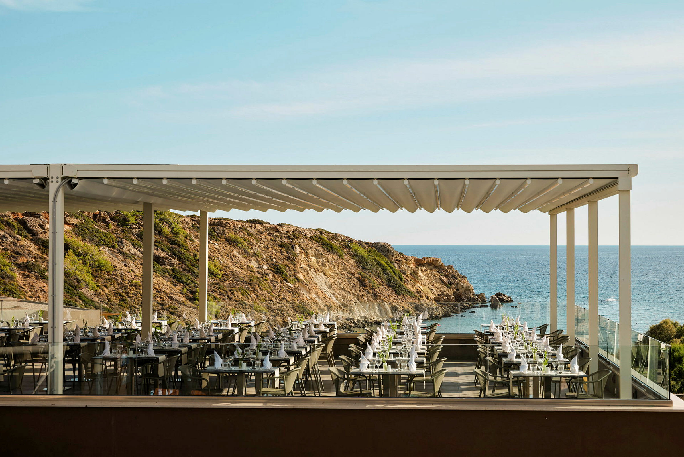 En utendørs restaurant med bord dekket og utsikt over havet og klipper. (AI-generert tekst)
