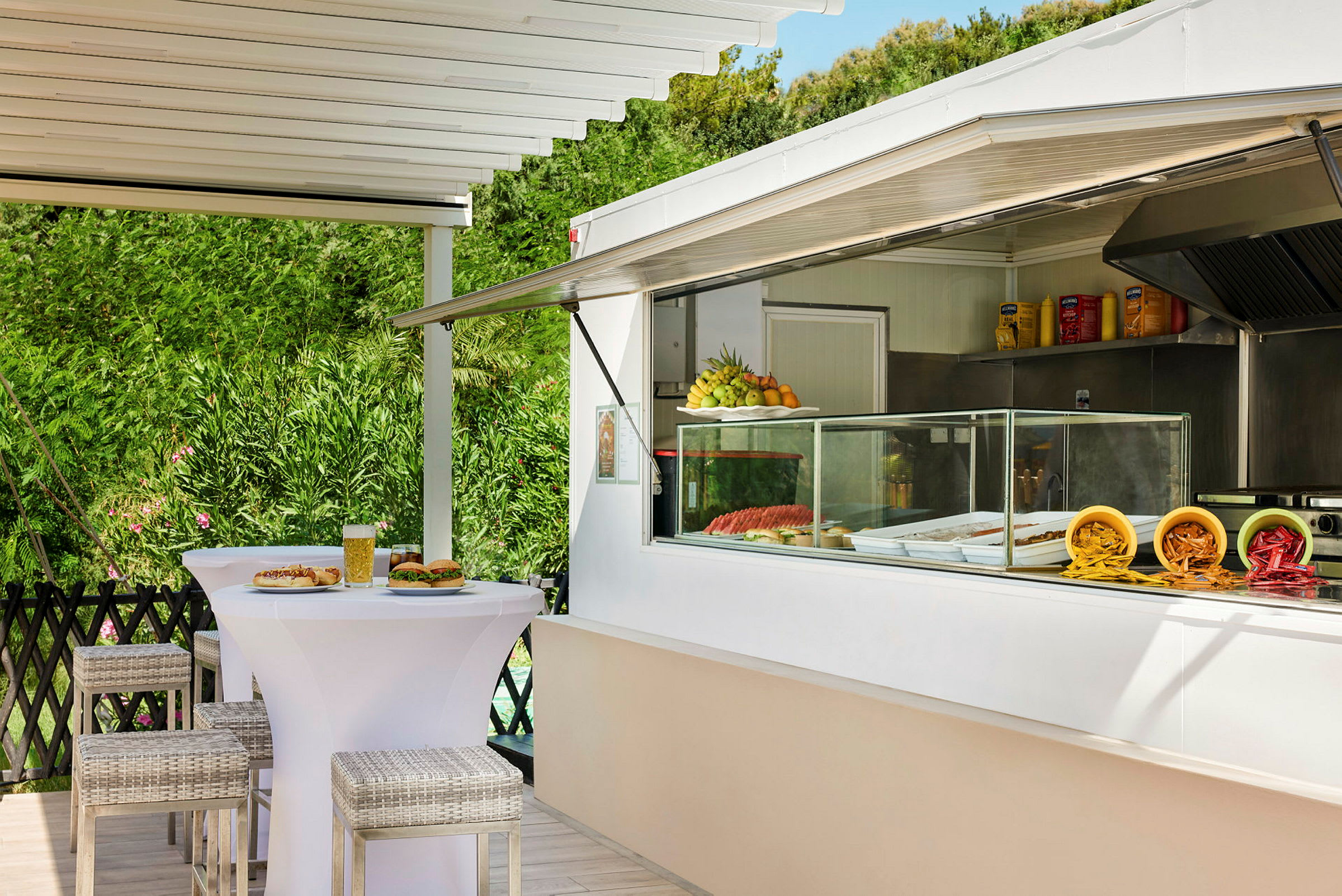 Bilde av en serveringsdisk ved en utendørsrestaurant med bord og stoler. På disken er tallerkener med mat og frukt, samt på bordet en øl og snacks. (AI-generert tekst)