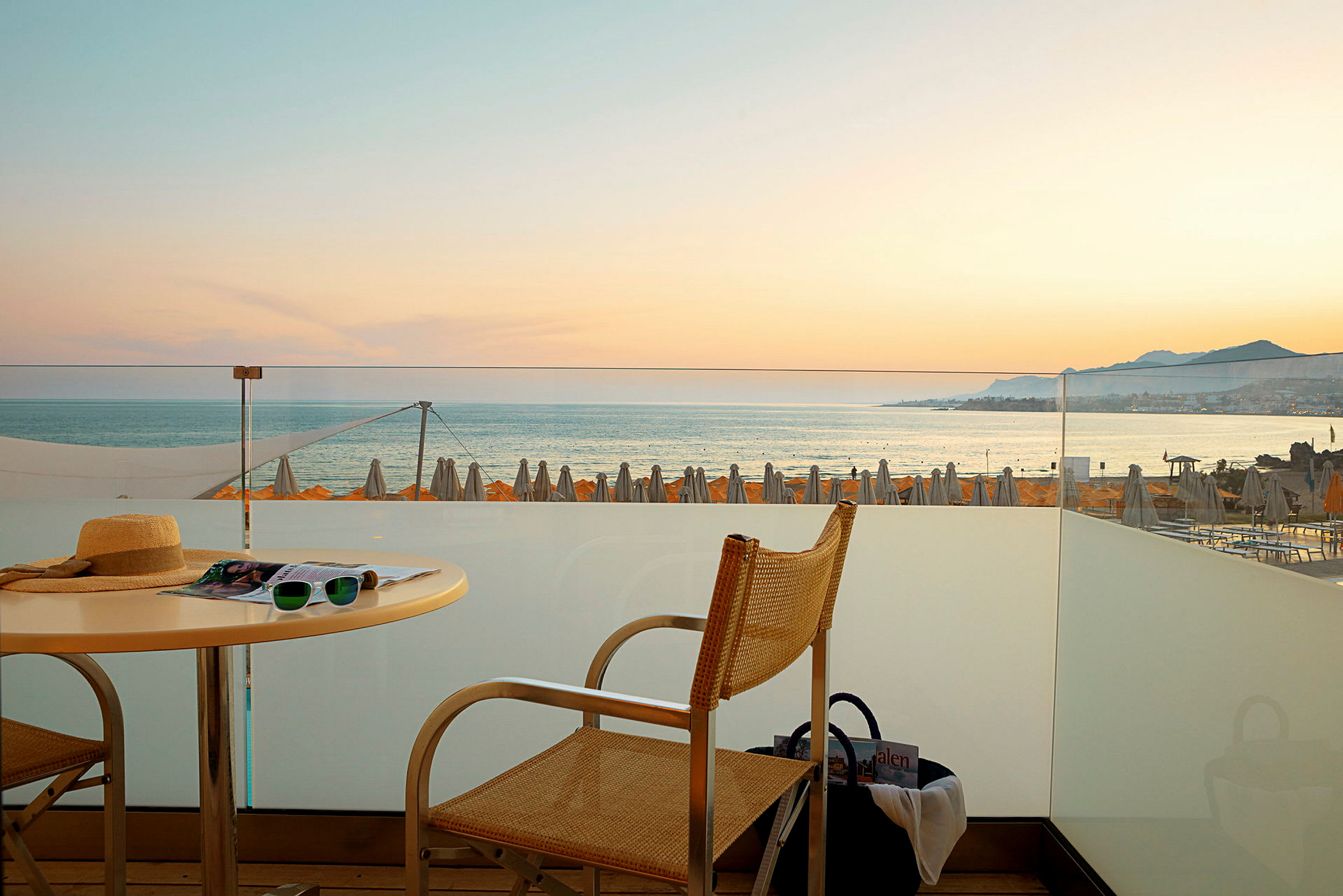Utsikt fra en terrasse mot havet med solnedgang og strandparasoller i bakgrunnen. (AI-generert tekst)