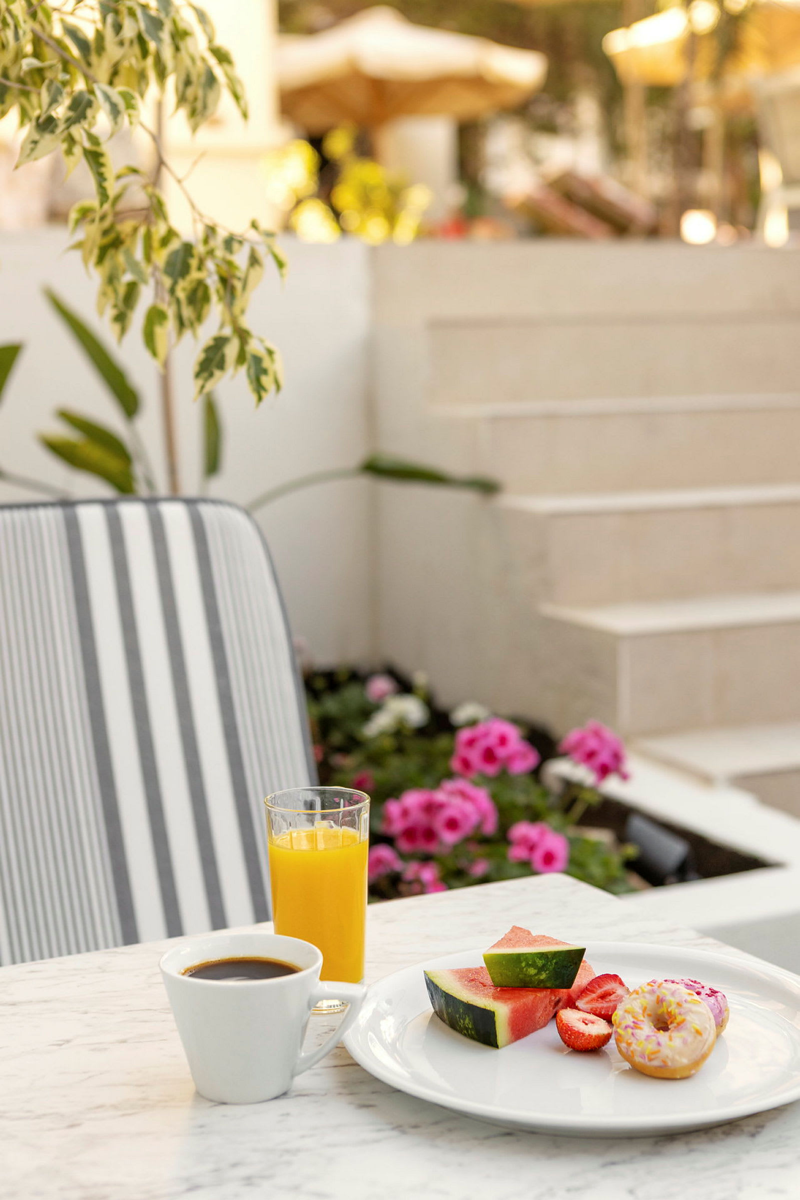 Et bord med en tallerken som inneholder vannmelonbiter, jordbær og en smultring ved siden av en kopp kaffe og et glass appelsinjuice. I bakgrunnen er blomster og trapper synlige. (AI-generert tekst)