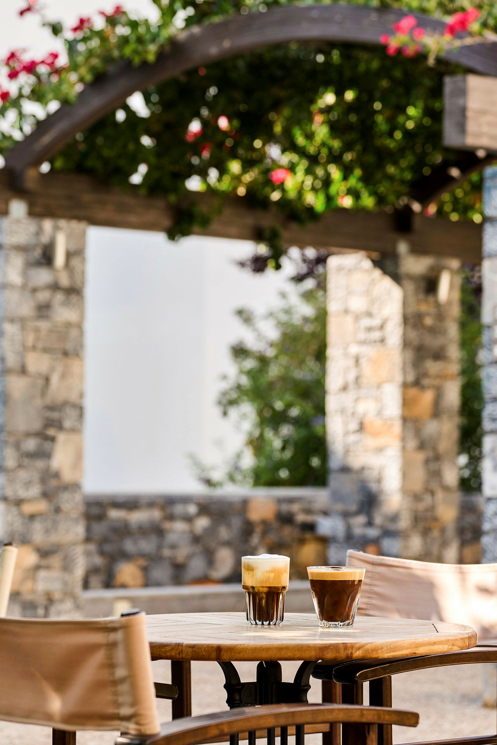 To kaffeglass står på et bord utendørs, med en steinstruktur og blomstrende planter i bakgrunnen. (AI-generert tekst)