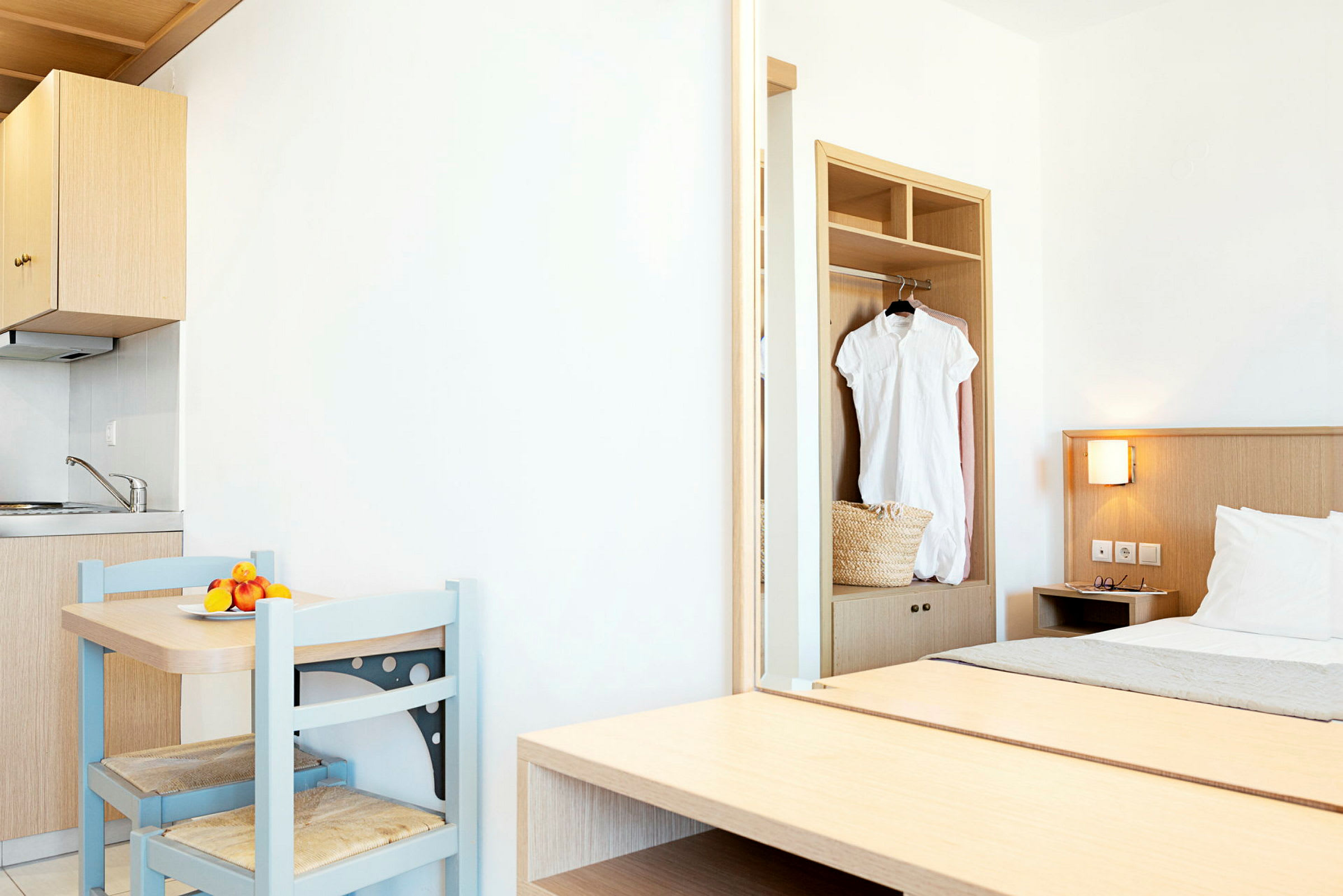 Et lyst hotellrom med en liten kjøkkenkrok, et bord med to stoler og en seng. En hvit kjole henger i skapet. (AI-generert tekst)