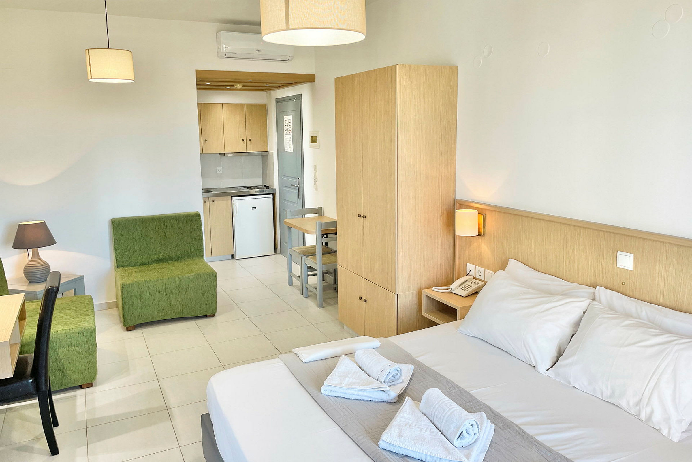 Et hotellrom med dobbeltseng, grønn sittegruppe, skrivebord, garderobe og en kjøkkenkrok. (AI-generert tekst)