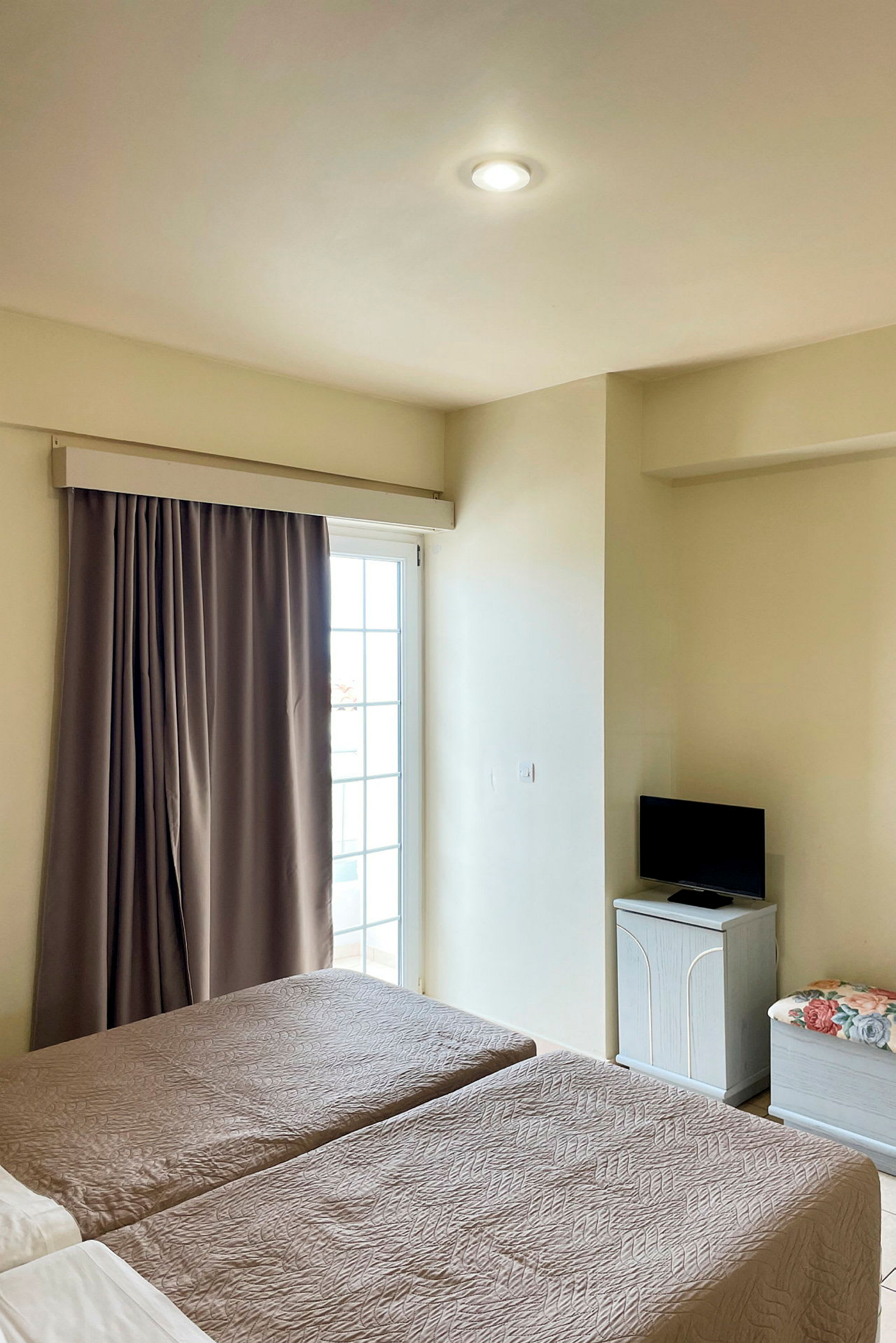 Et hotellrom med to enkeltsenger, en balkongdør med gardin og en liten TV på et skap. (AI-generert tekst)