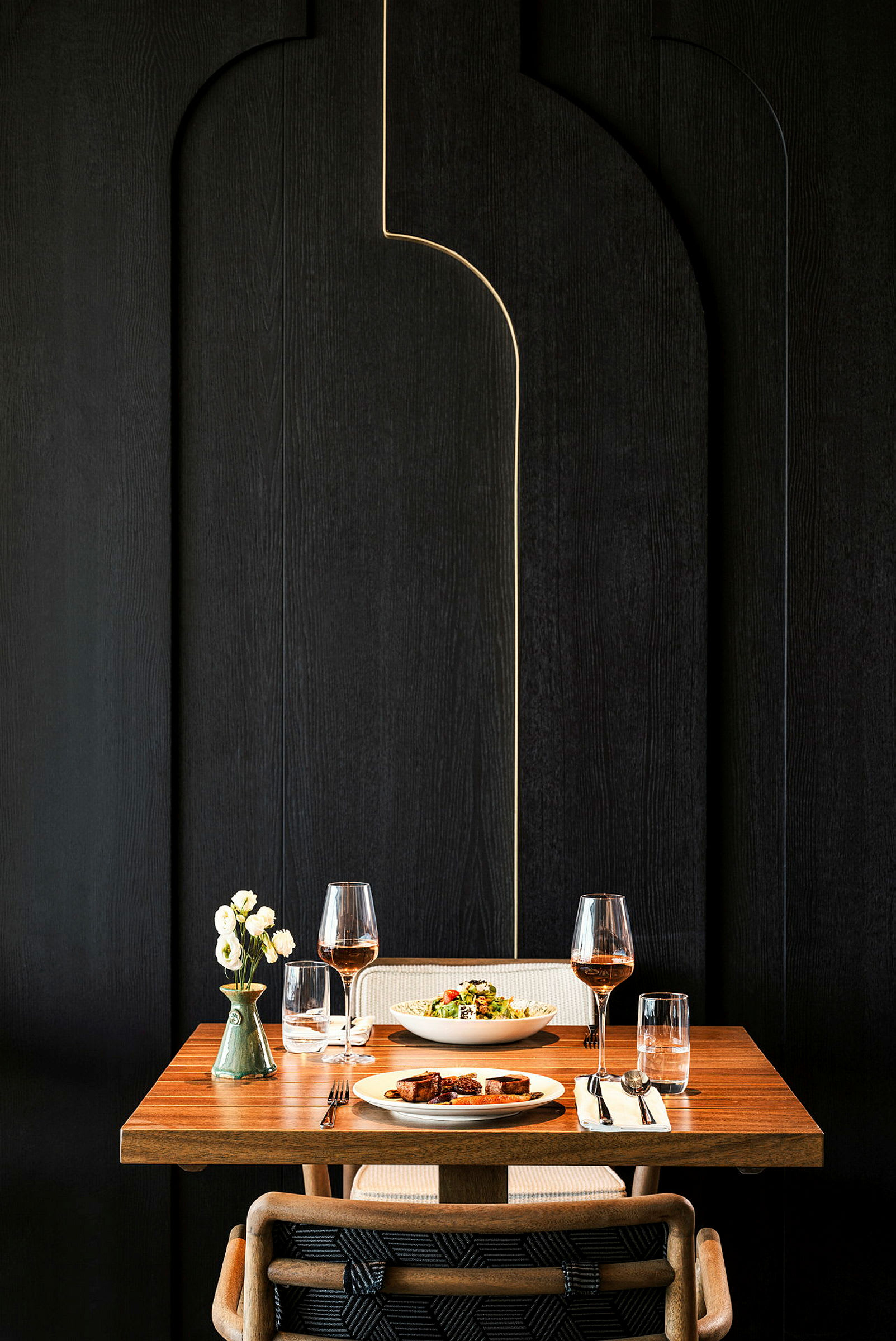 Et dekket bord med en overflate av tre, for to personer, to glass vin, to tallerkener med mat og en vase med blomster. (AI-generert tekst)