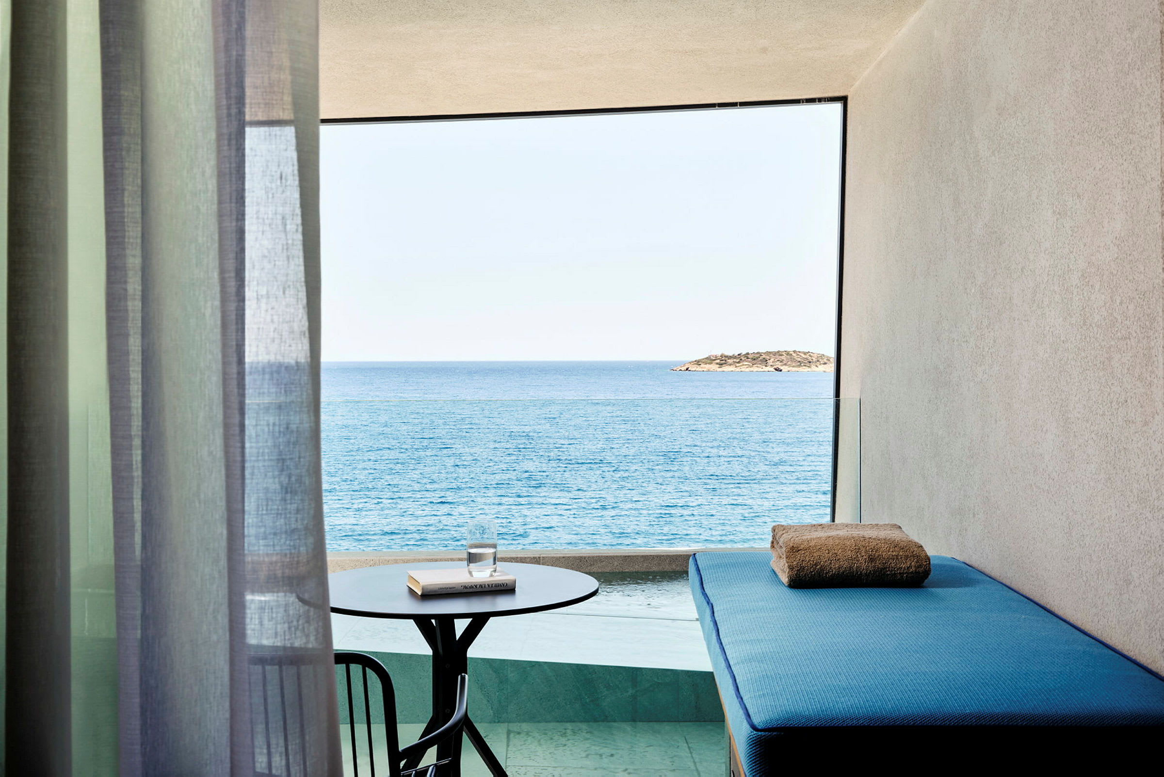 En balkong med utsikt over havet og en øy i det fjerne. På balkongen står det et lite bord med et glass vann og en bok samt en blå solseng med et håndkle. (AI-generert tekst)