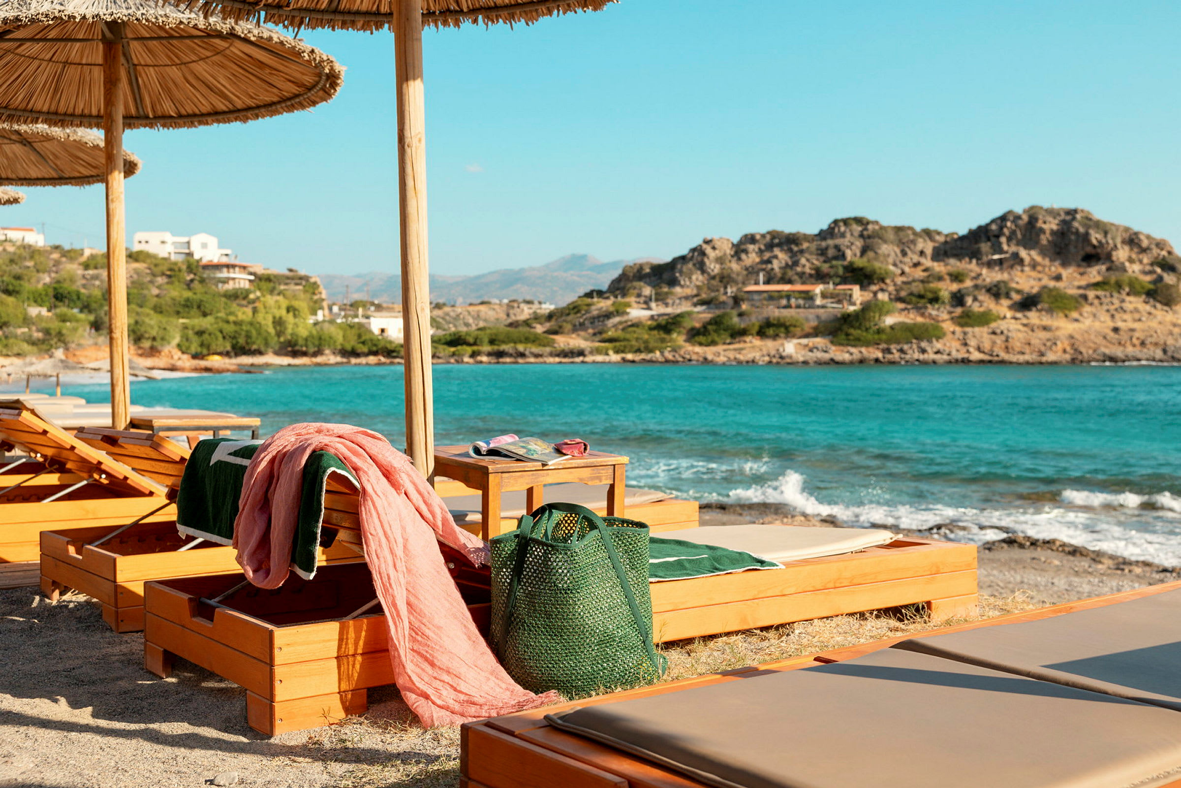 Solstoler med tøydekte puter og parasoller står på en strand med utsikt over et turkis hav og steinete kyst. (AI-generert tekst)
