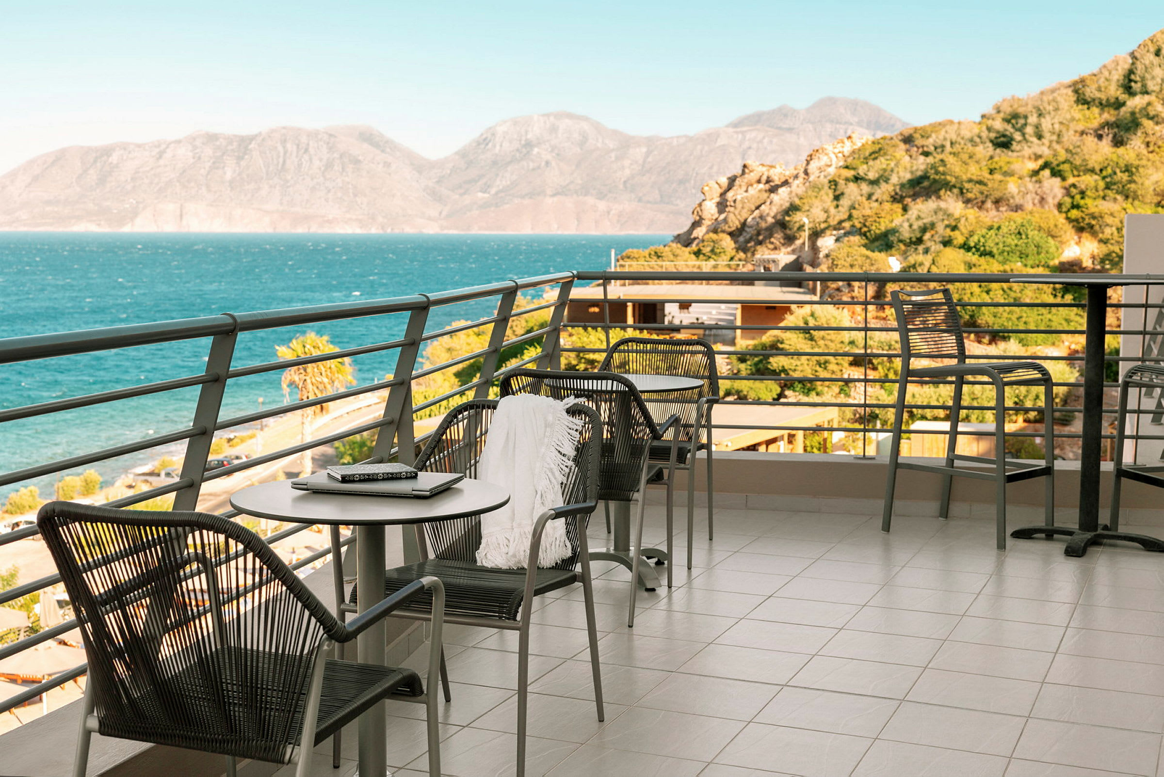 En terrasse med bord og stoler, omgitt av et rekkverk. Utsikt over havet og fjellene i det fjerne. (AI-generert tekst)