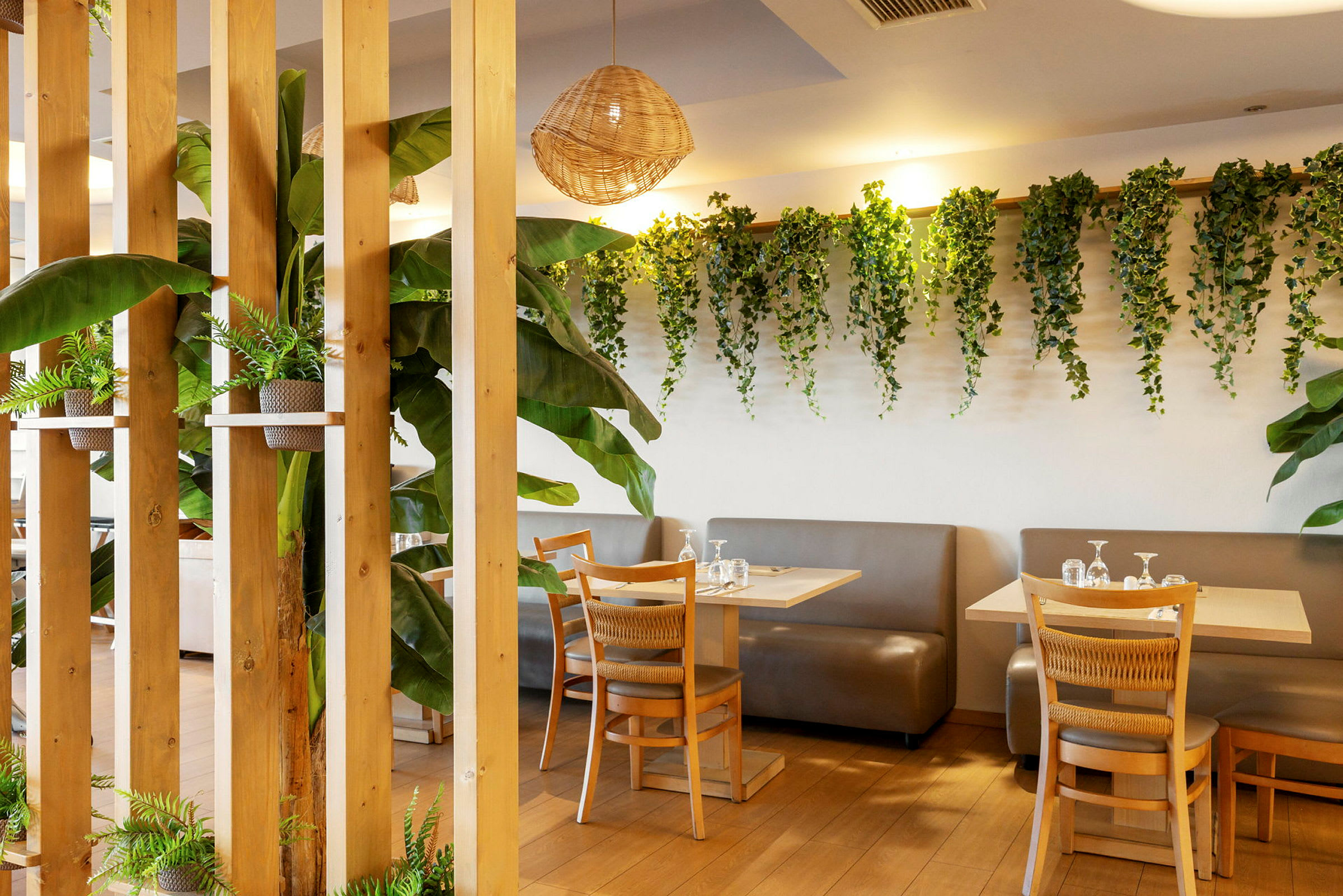 En innendørs restaurant med bord og stoler i tre. Veggene er dekorert med grønne planter, og det finnes betydelige innslag av grønne planter rundt om. (AI-generert tekst)