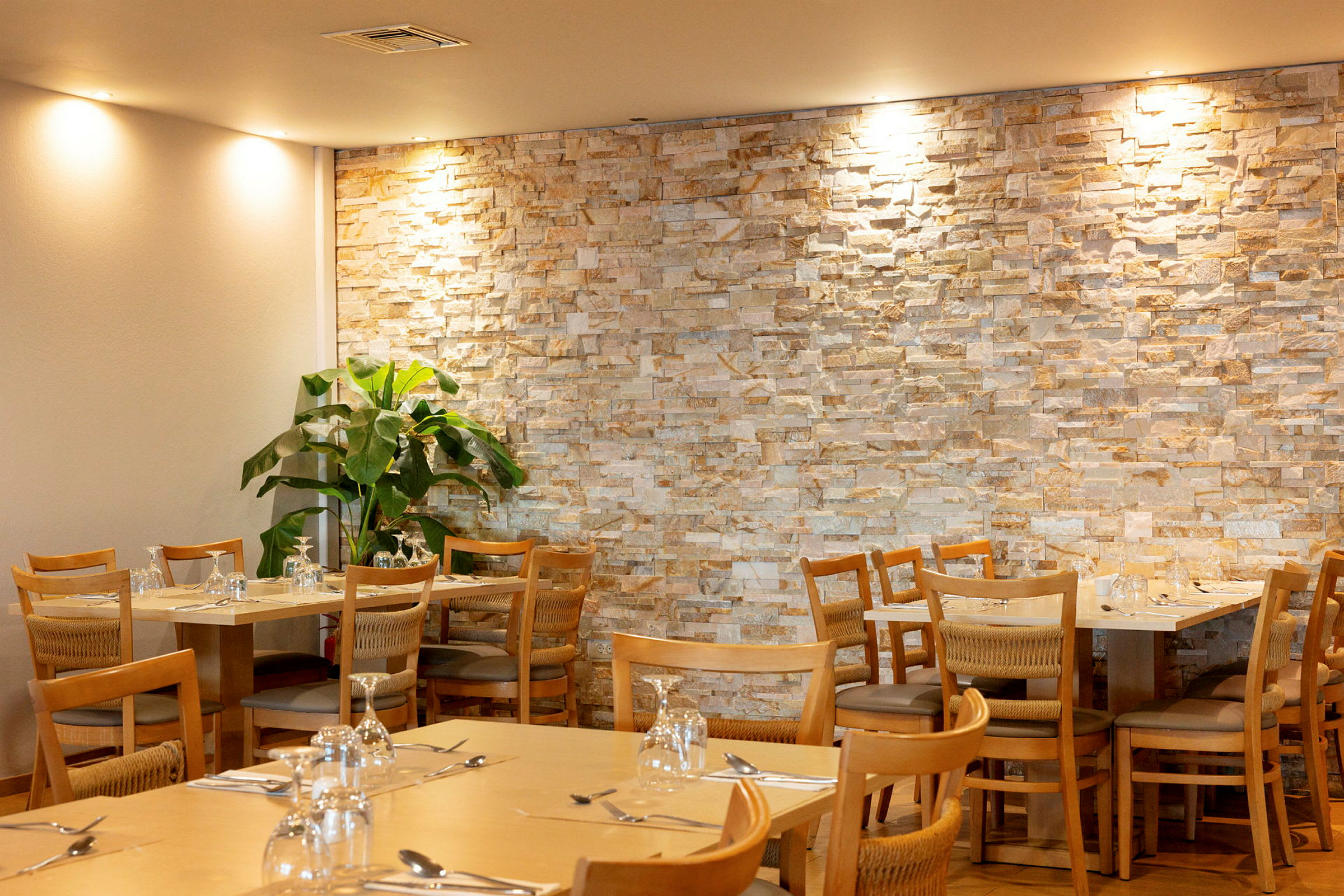 Restaurantmiljø med bord og stoler foran en vegg av stein. En grønn plante står i hjørnet. (AI-generert tekst)