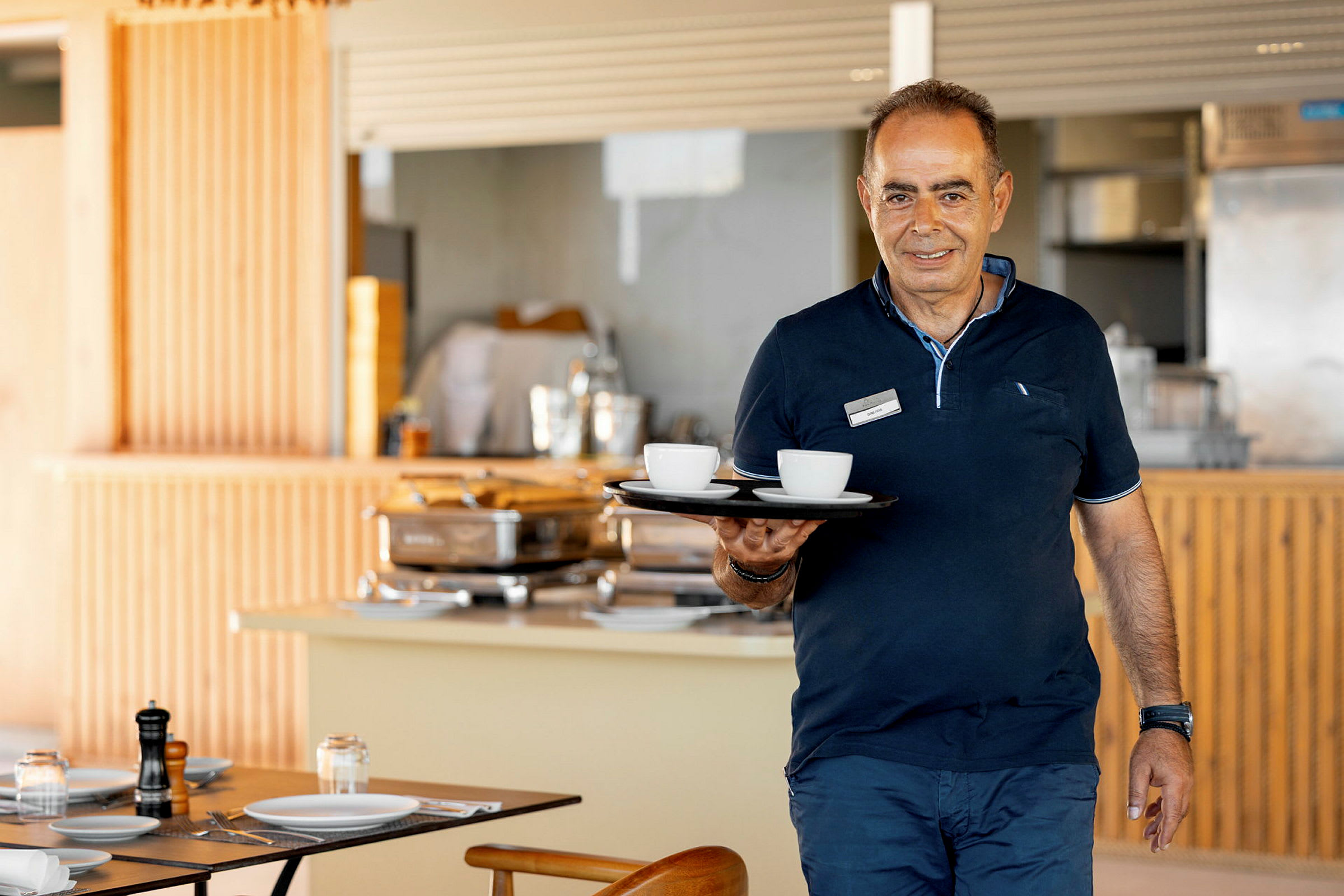 En person holder et brett med kaffekopper i en spisesal. (AI-generert tekst)