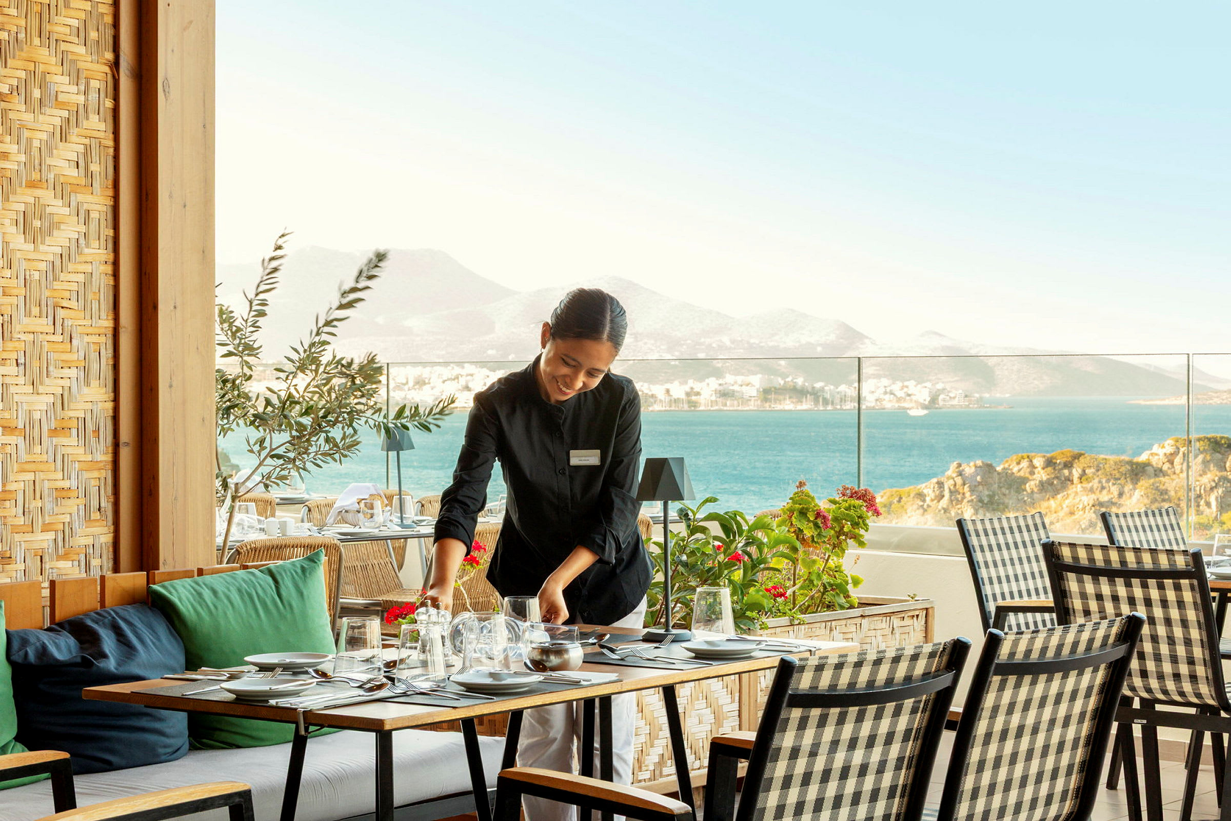 En servitør dekker et bord på en terrasse med havutsikt og fjell i bakgrunnen. (AI-generert tekst)