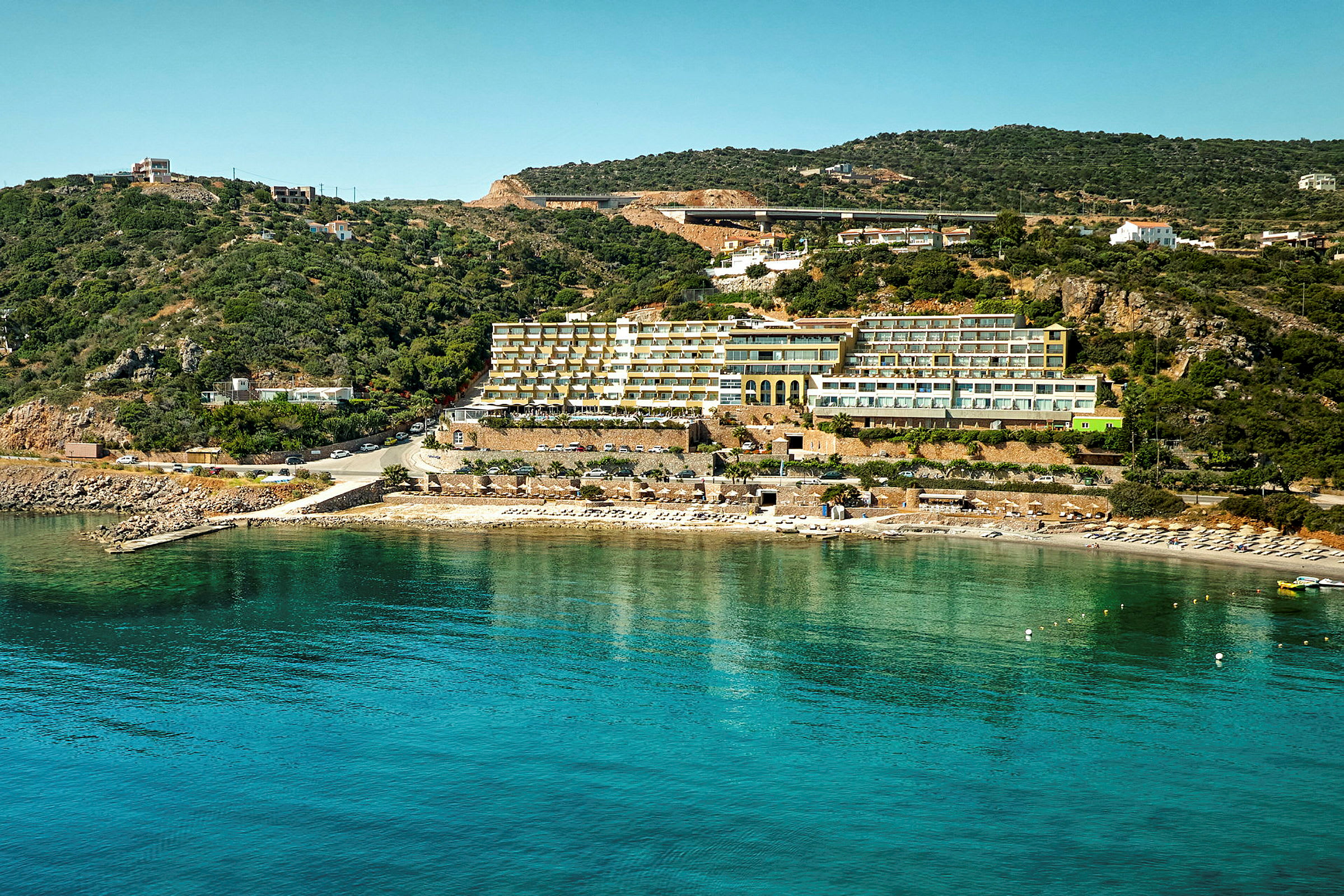 Hotell ved strand med klart blått vann og grønne åser i bakgrunnen. (AI-generert tekst)