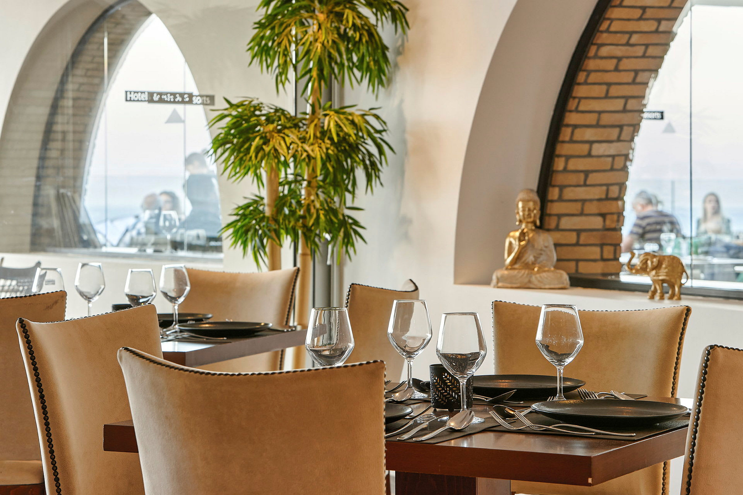 Et restaurantbord med oppdekt servise og glass, omgitt av elegant interiør med grønne planter og dekorative figurer i bakgrunnen. (AI-generert tekst)