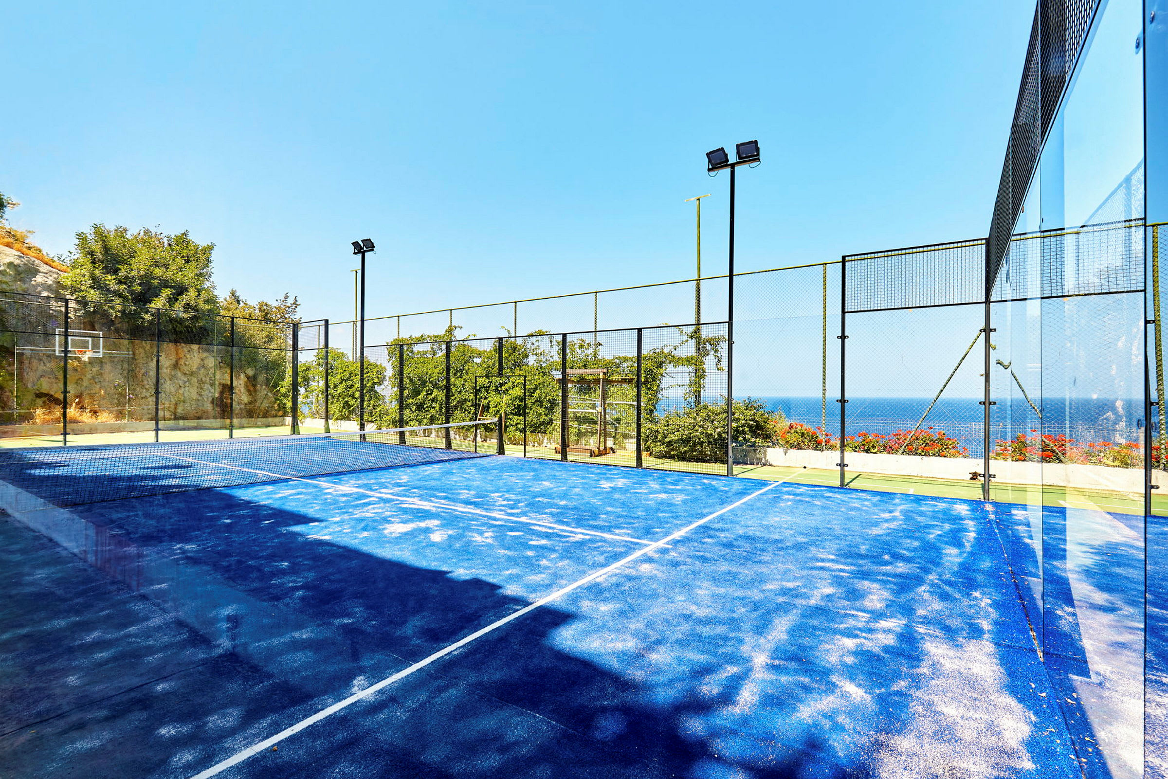 En padelbane med blått underlag omgitt av gjerder med utsikt over havet. (AI-generert tekst)